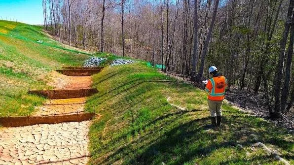 A person wearing an orange safety vest and white helmet standing on a grassy slope near a forest, with an area of cracked dry earth and erosion control structures in the foreground.