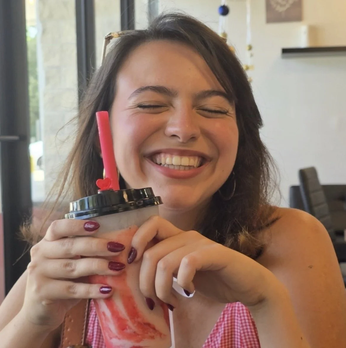 A woman with shoulder-length brown hair and a checkered pink dress smiling with closed eyes while holding a drink with a pink straw.