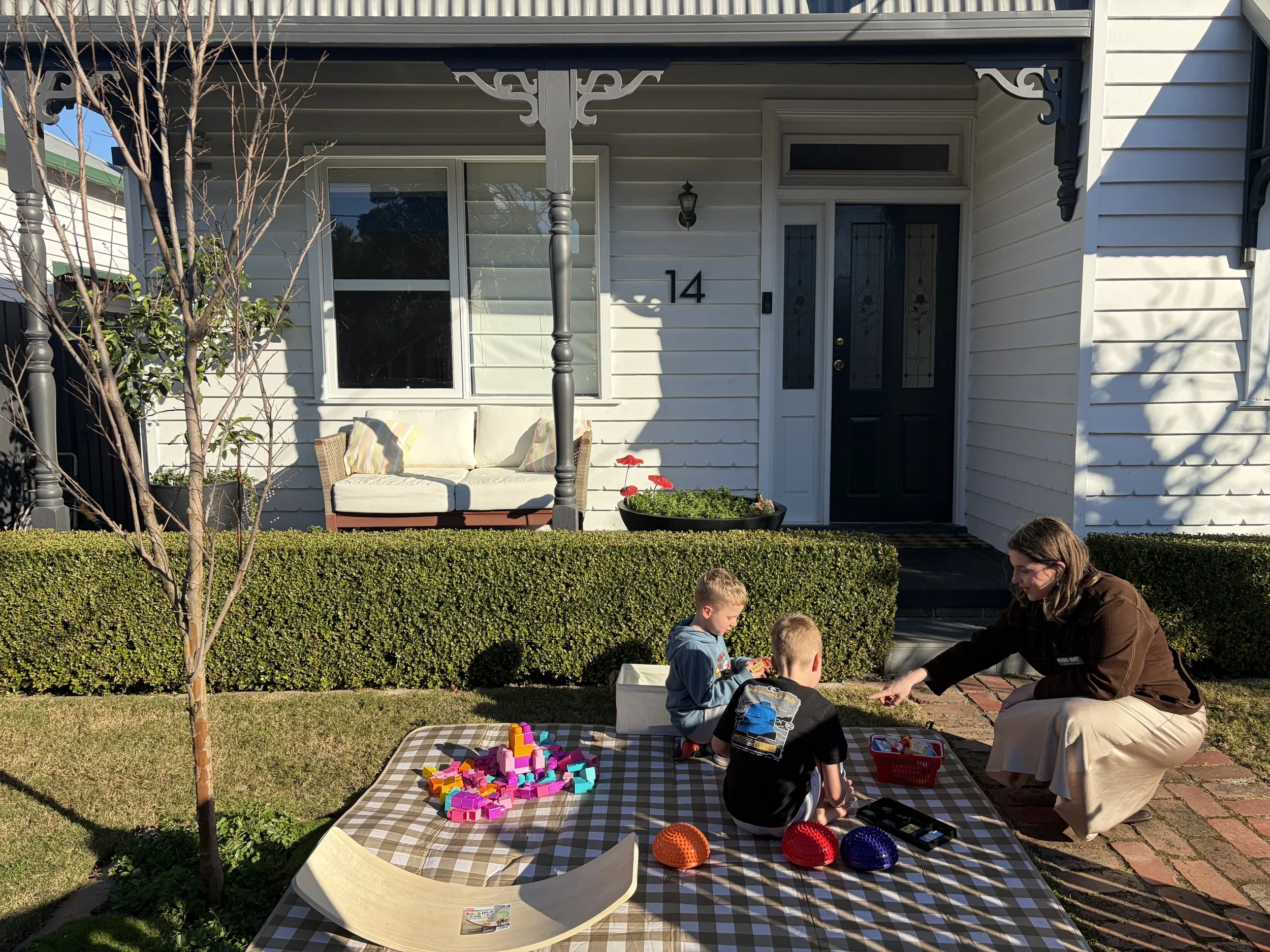 A woman and three children playing with colorful plastic blocks on a checkered tablecloth outside a house during the daytime.