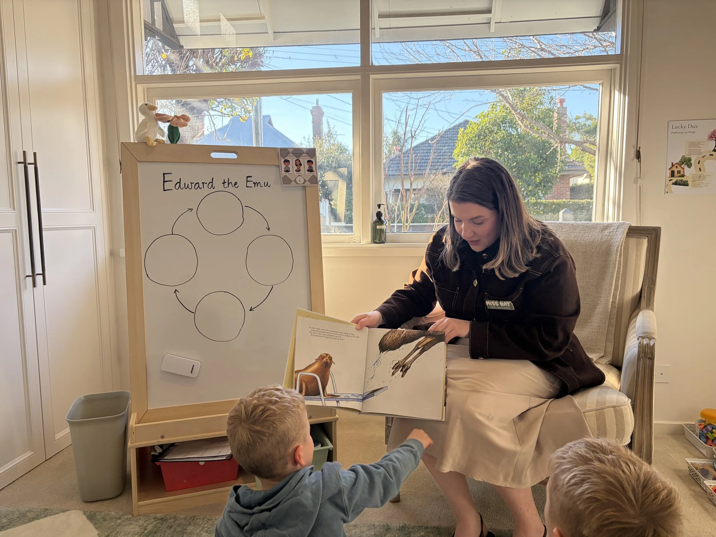 A woman sitting on her knees, reading a children's book to a group of young children who are sitting on the floor, in a well-lit room with a large window showing trees and rooftops outside. There is a whiteboard with a diagram of a bird and the title 'Edward the Emu' in front of her, and some toys and a trash bin in the room.