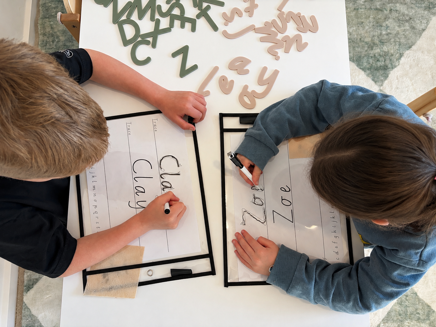 Two children are writing on valentine's day themed posters with black markers; one poster says "I love" with a picture of a cat, the other says "Zoe".