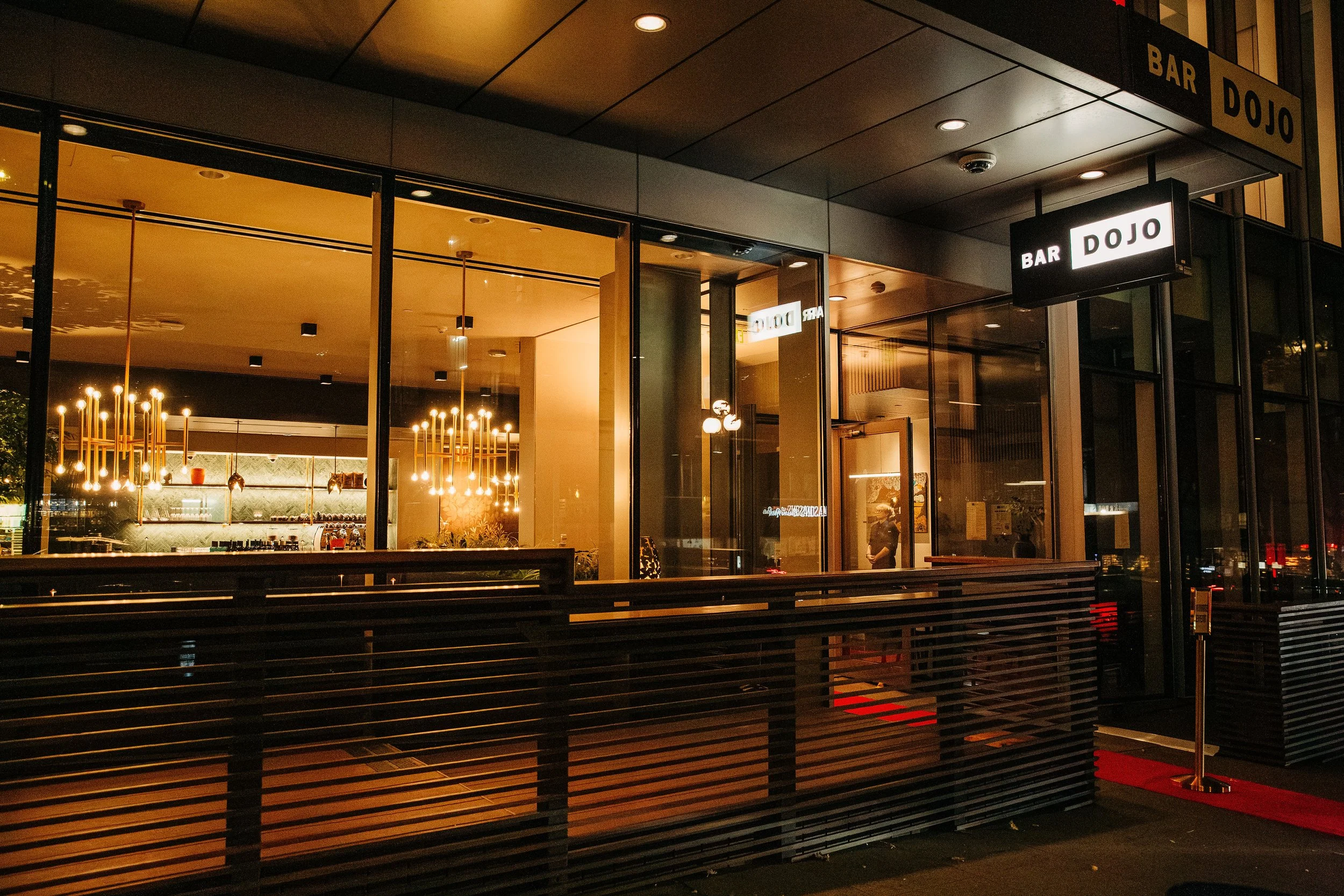 Interior of a modern cafe with patrons and staff, featuring wooden flooring, a barista station with a coffee machine, and a large decorative wall with gold floral designs.