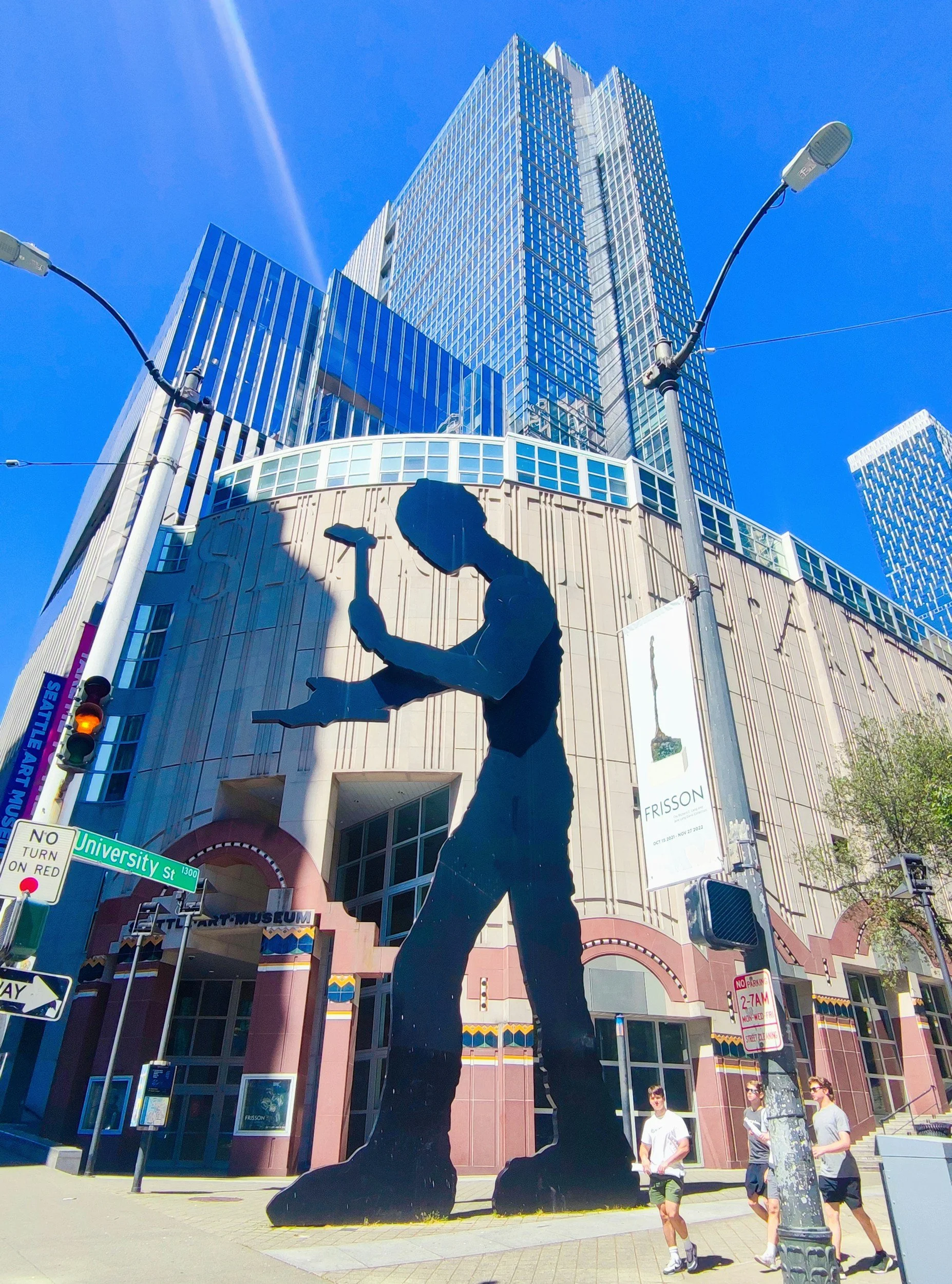 A large skyscraper building with glass windows against a blue sky, with a black silhouette sculpture of a person standing on their knees, holding an object, in the foreground. There are street signs, light poles, and people walking nearby.