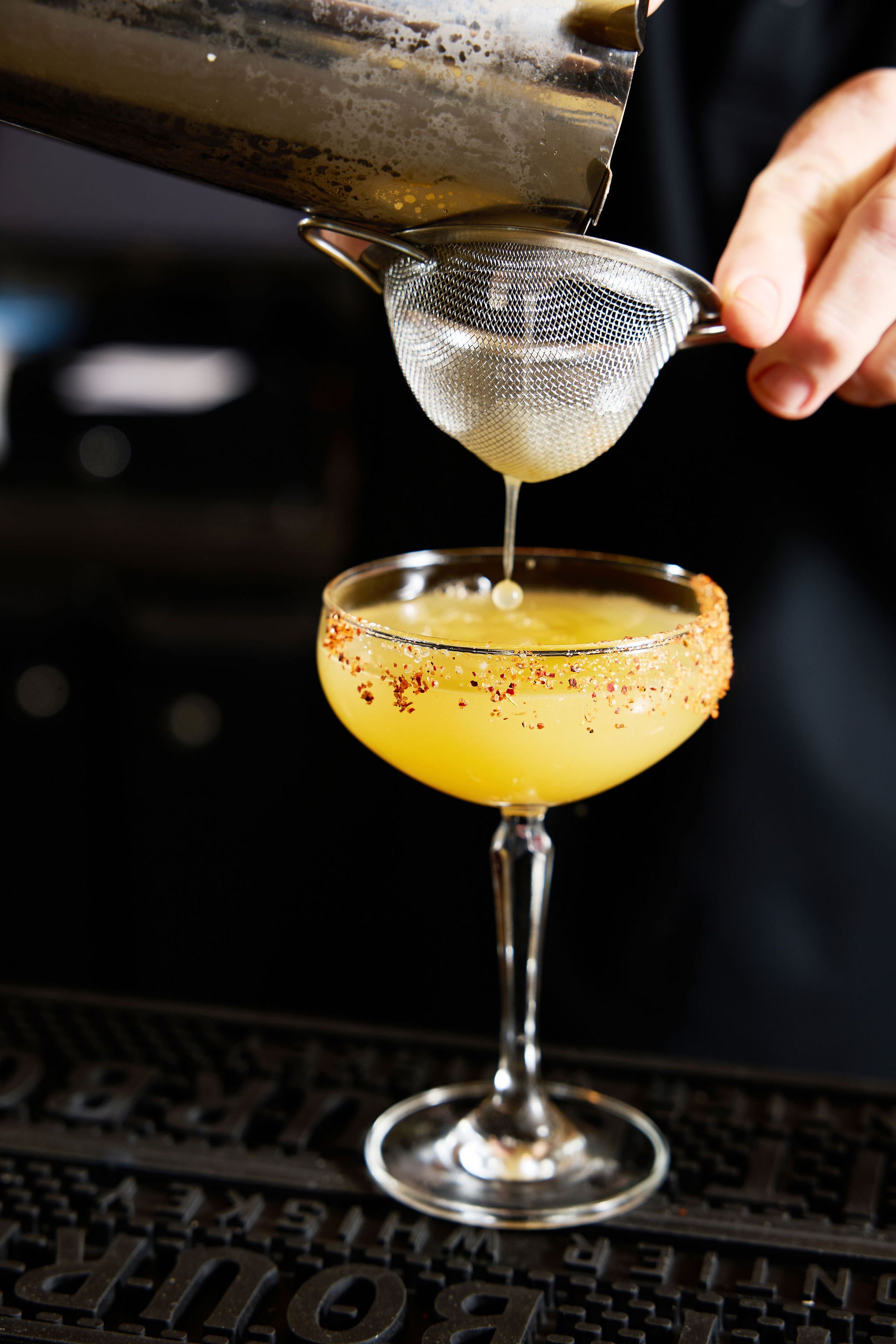 A cocktail glass with a salted rim, containing a yellow drink, sits on a black bar mat. A strainer is held above the glass, pouring a liquid into it. The background is dark and blurred.