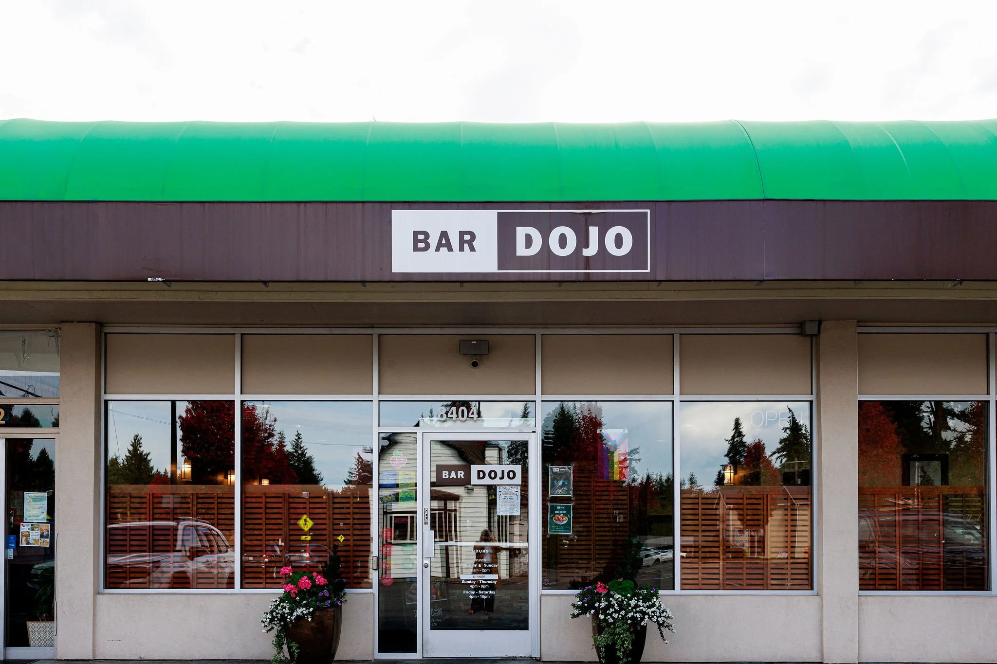Front view of a building with a sign reading 'BAR DOJO' above the entrance, glass doors, and windows; two large flower pots with colorful flowers on either side of the entrance, and reflections of trees and buildings in the windows.