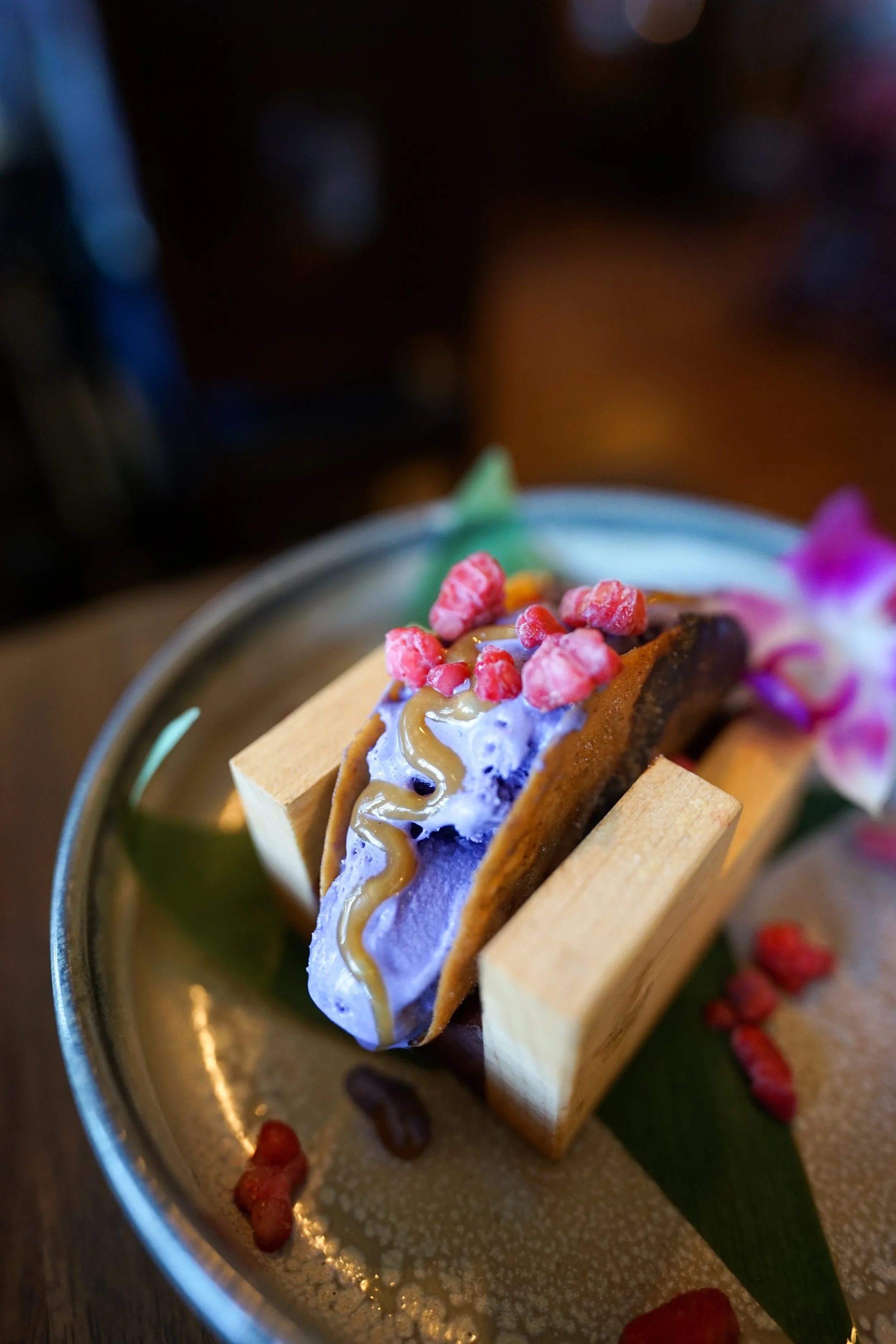 A gourmet ice cream dessert topped with caramel sauce, red berries, and purple flower petals on a small round dish.