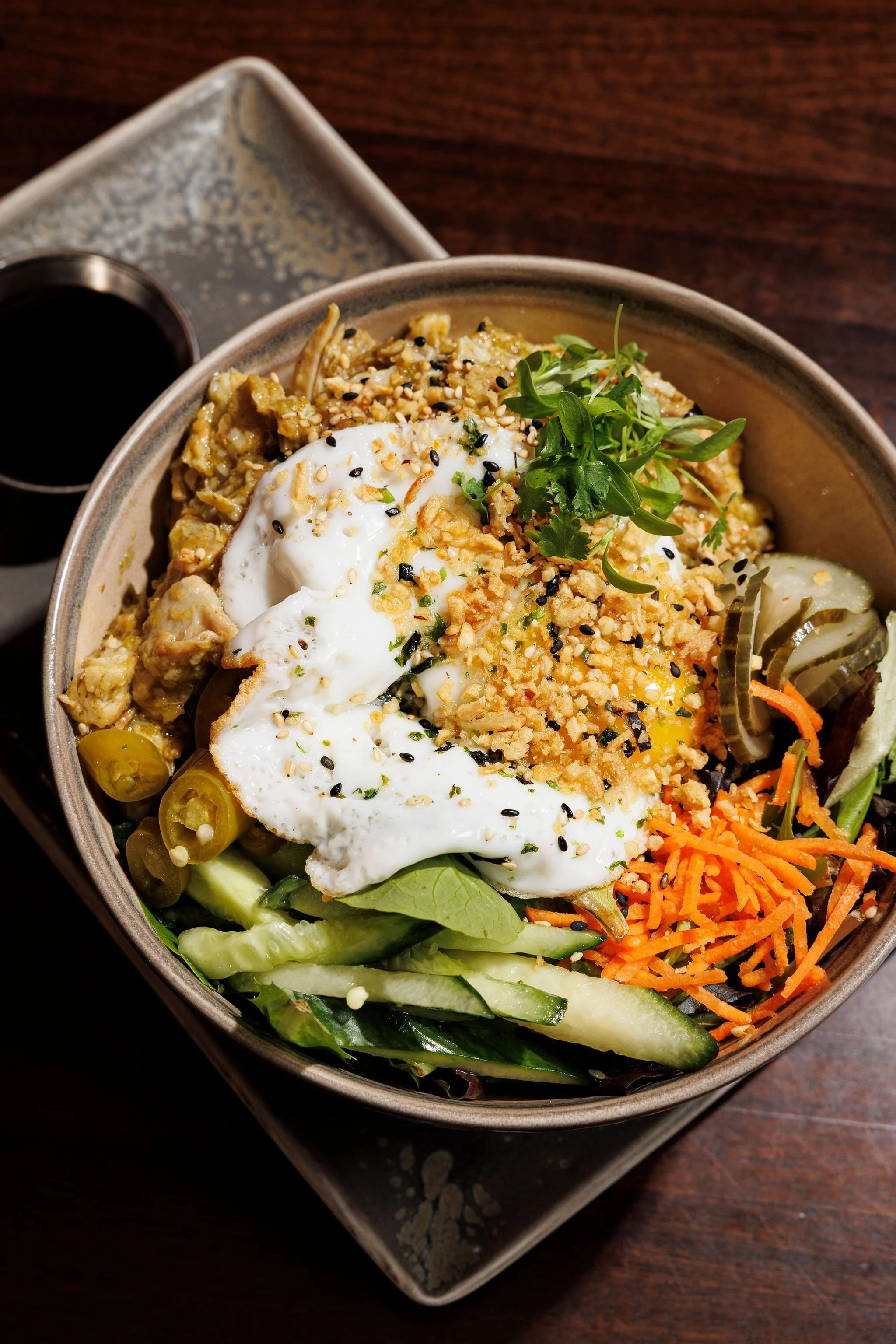 Buddha bowl with rice, shredded carrots, sliced cucumber, green salad, topped with a poached egg, microgreens, chopped nuts, and black sesame seeds, with soy sauce on a side dish.