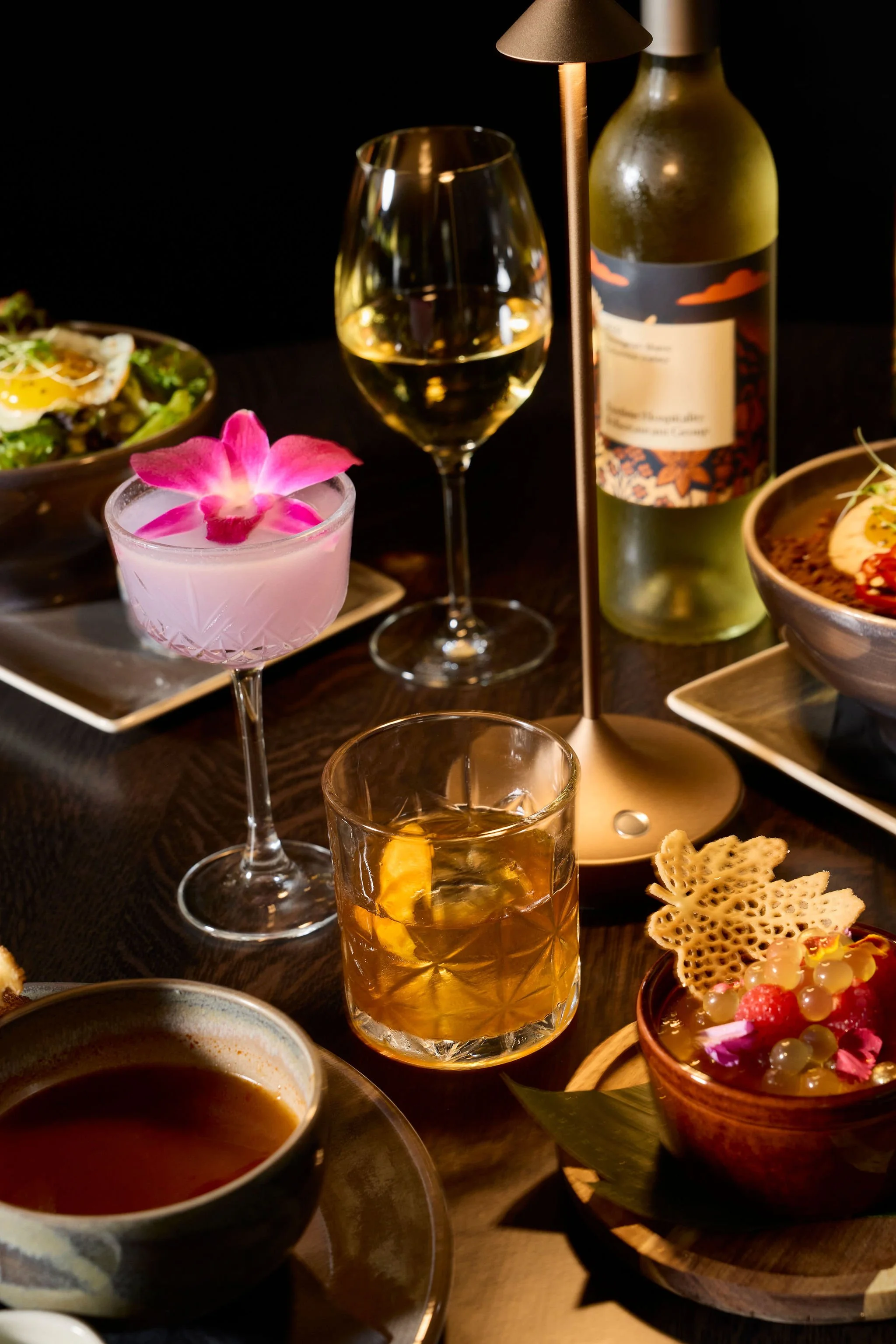 Elegant cocktail table with whiskey, wine, and pink flower cocktails, a wine bottle, lamp, and assorted gourmet dishes.