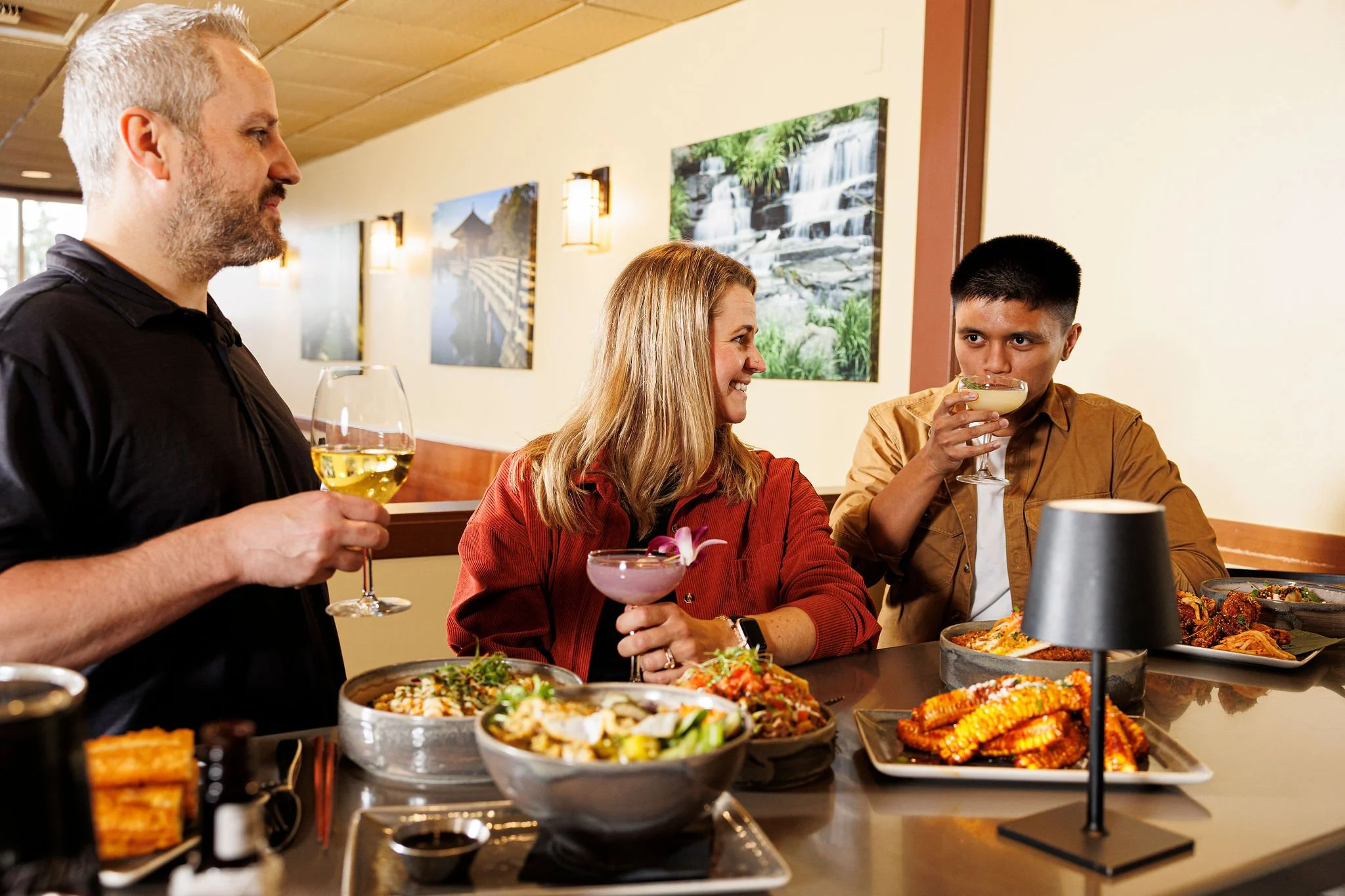 A group of three people enjoying a meal at a restaurant, with various dishes and drinks on the table, including wine and cocktails, and framed nature pictures on the wall.