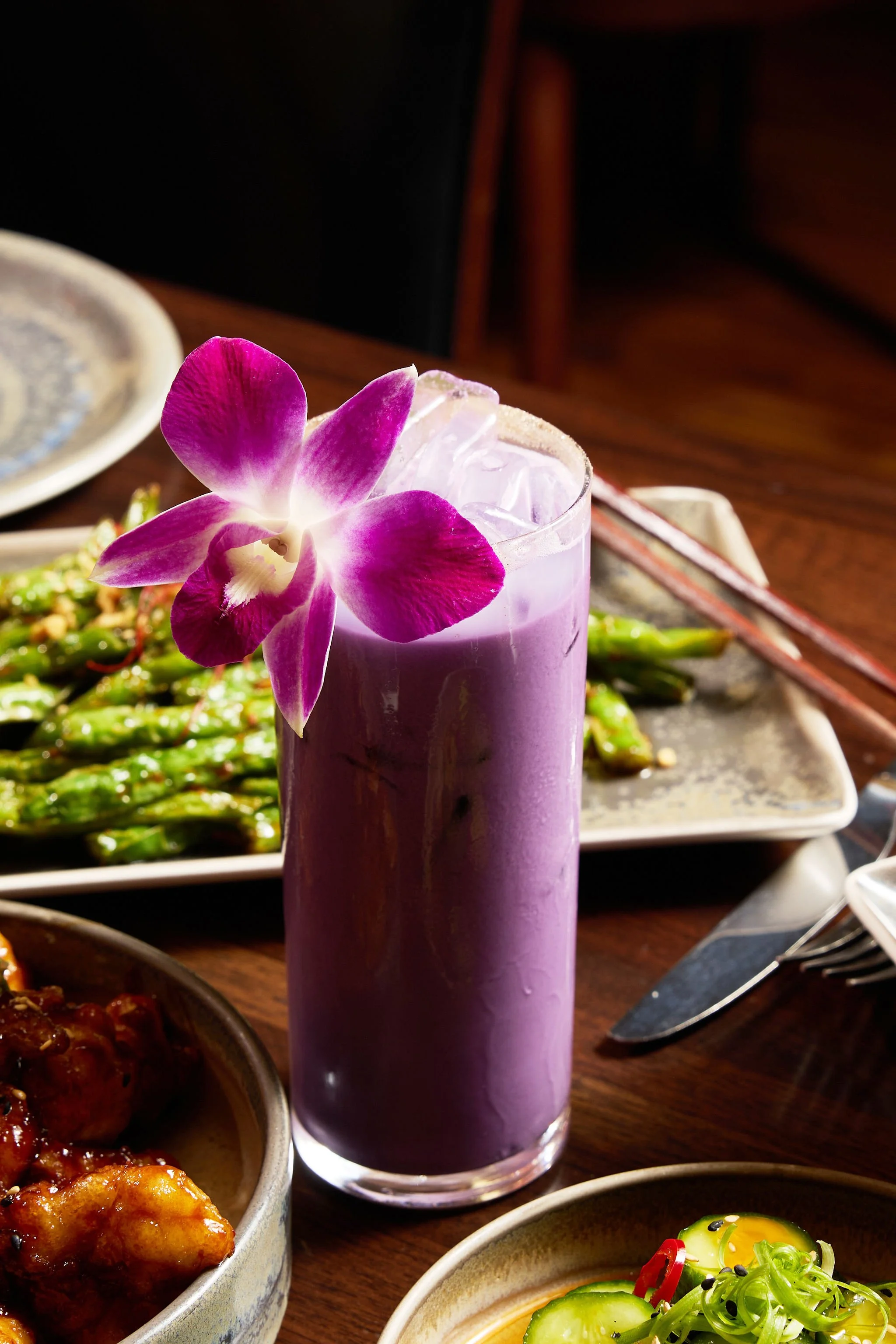 A purple fondant cake decorated with a pink and white orchid flower on top, surrounded by various Asian dishes including vegetables and sauces on a wooden table.