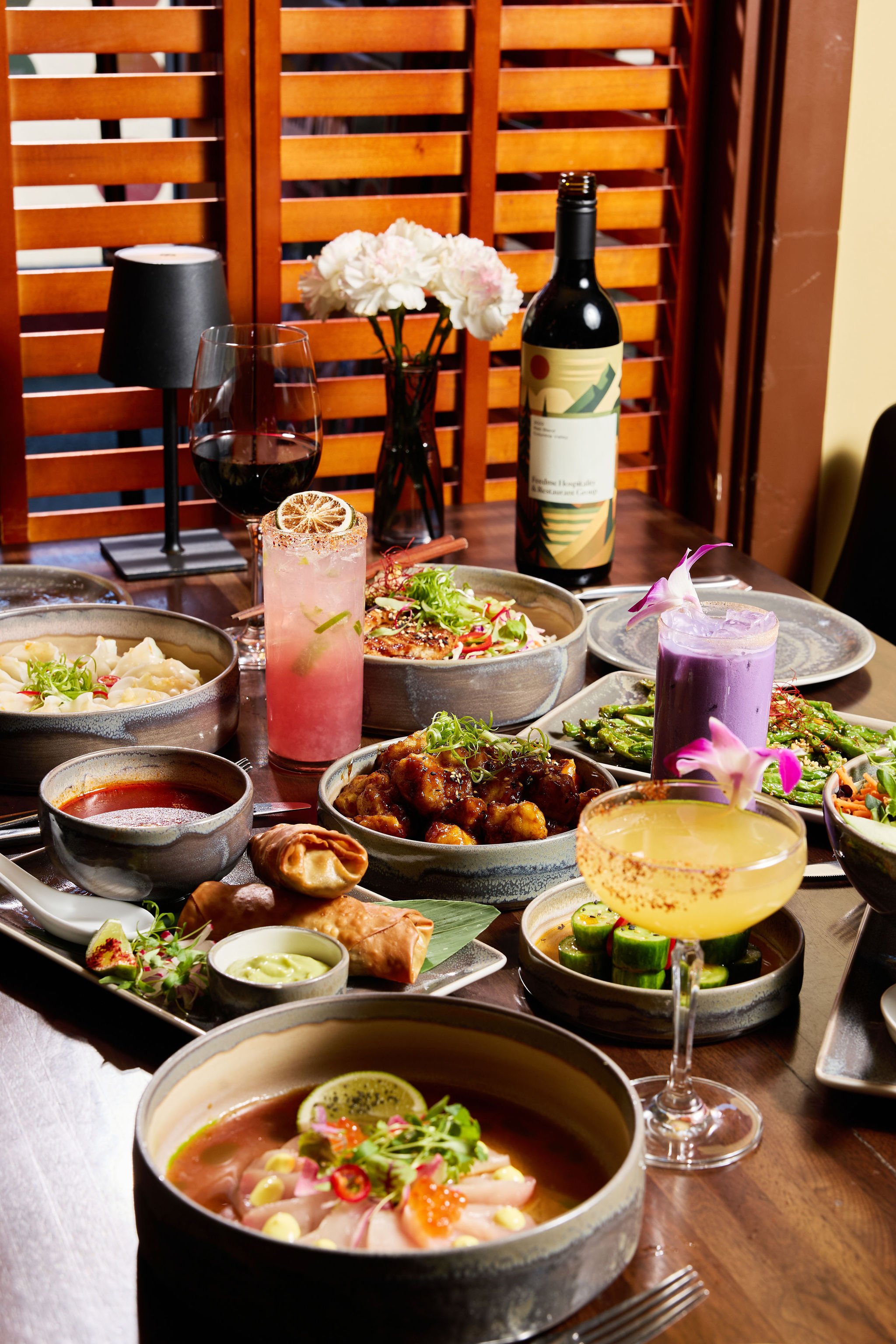 A dining table set with various dishes including sashimi, chicken dish, cucumber salad, asparagus, and multiple cocktails, with a bottle of wine, a vase of flowers, and black table lamps in the background.