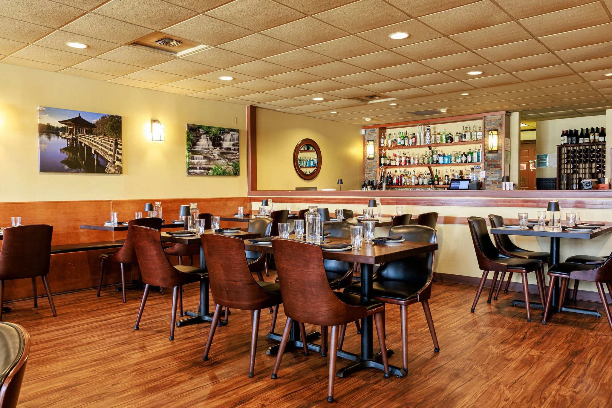Interior of a restaurant dining area with wooden floors, dark wood tables, and chairs. The wall has framed pictures of nature scenes, and a bar area with bottles of alcohol and a mirror is visible in the background.