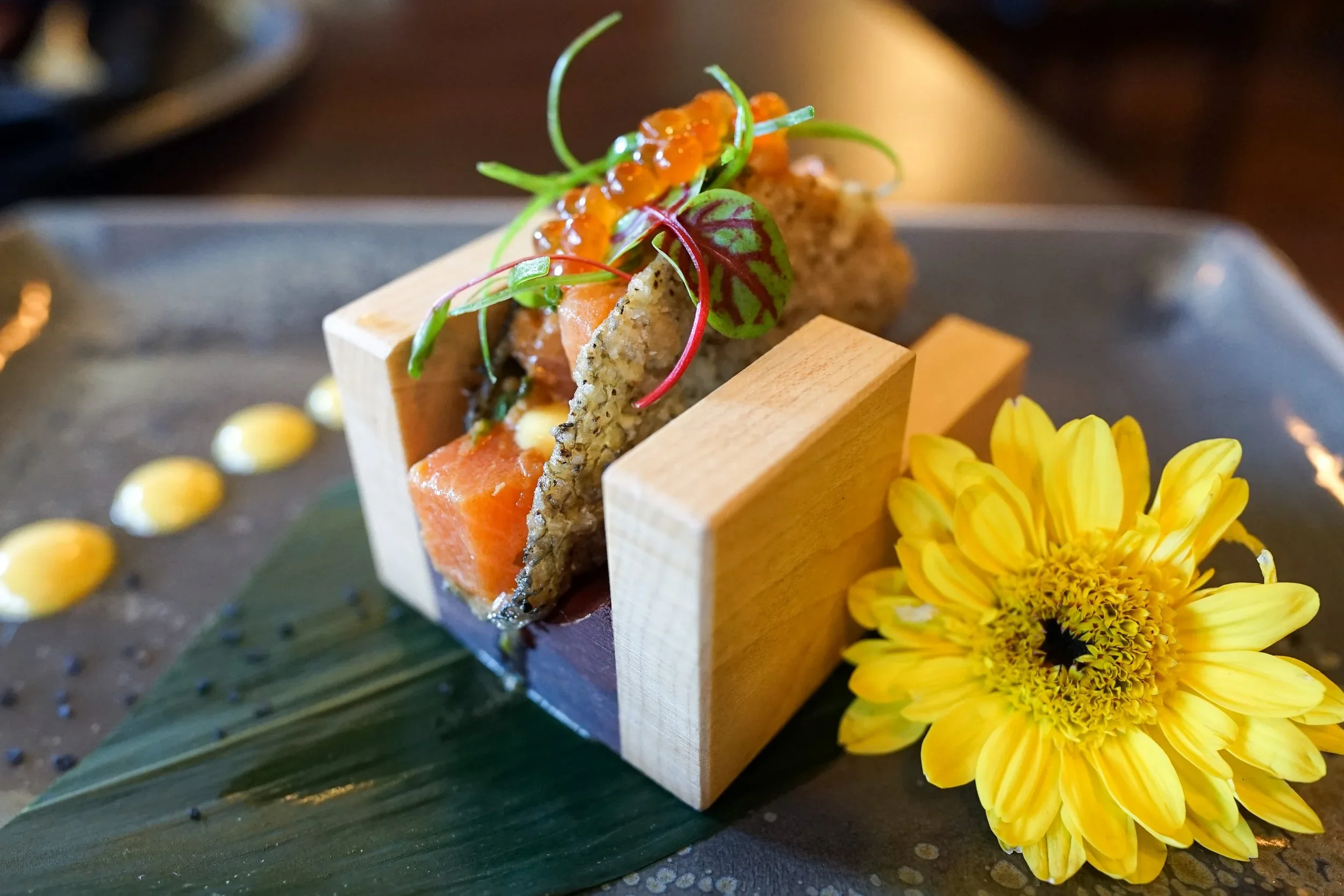 A plated appetizer featuring smoked salmon, microgreens, and caviar on crispy bread, garnished with yellow flowers and serving as a culinary presentation.