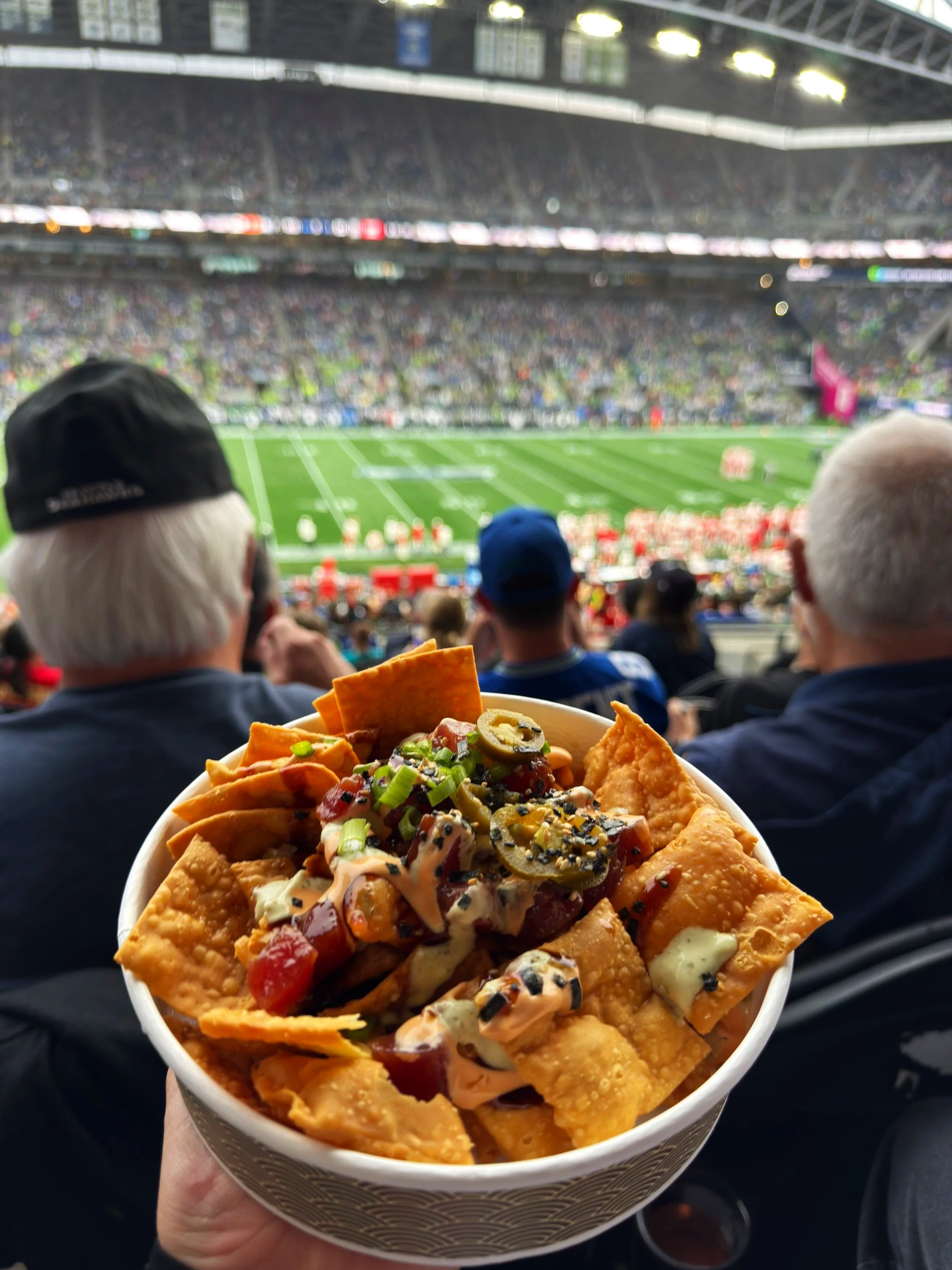 A bowl of nachos with cheese, toppings, and guacamole held up in front of a crowded football stadium with fans and players on the field.