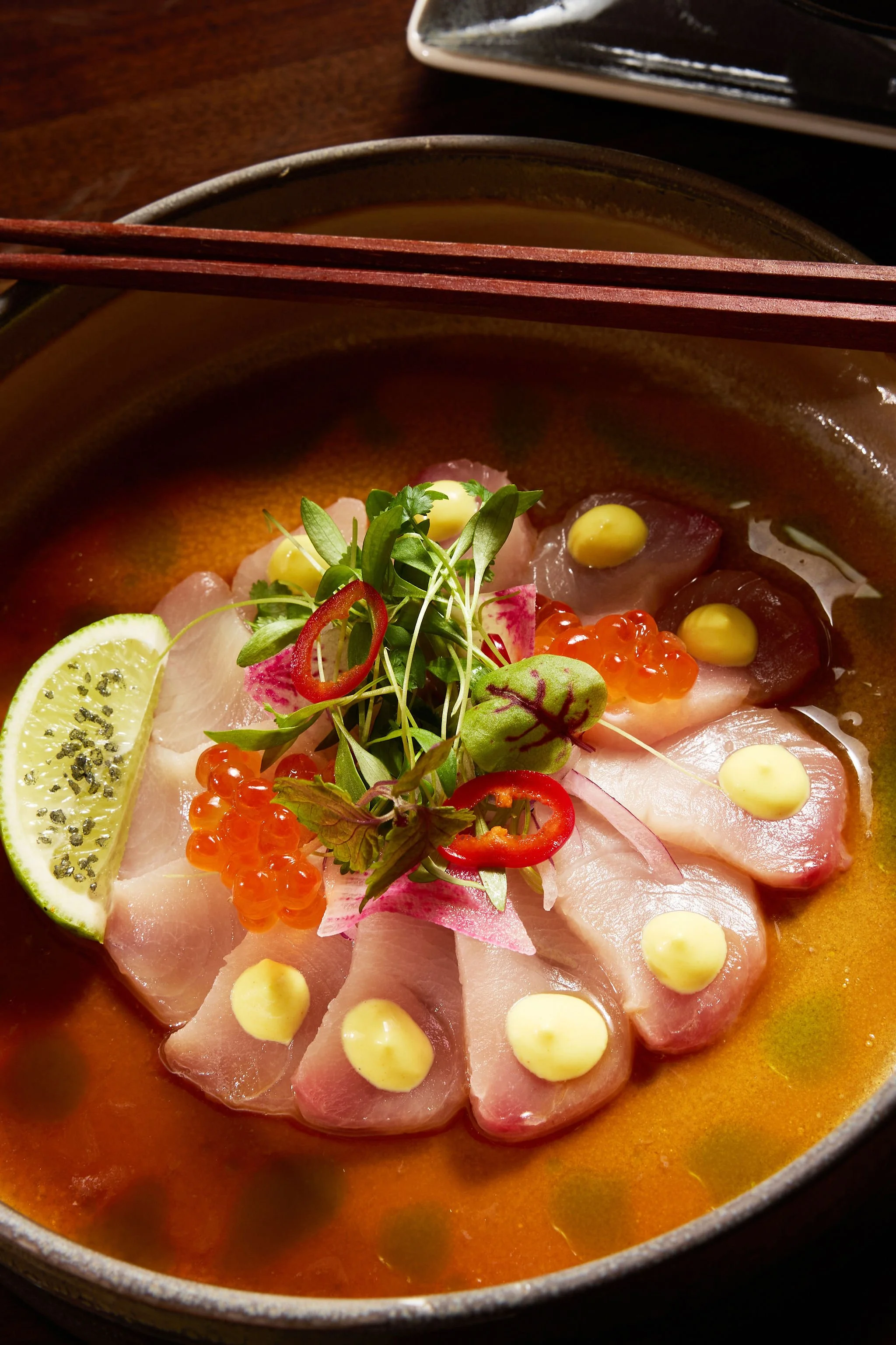 Close-up of a bowl of sashimi garnished with lemon, microgreens, and fish roe, with chopsticks resting on the bowl.
