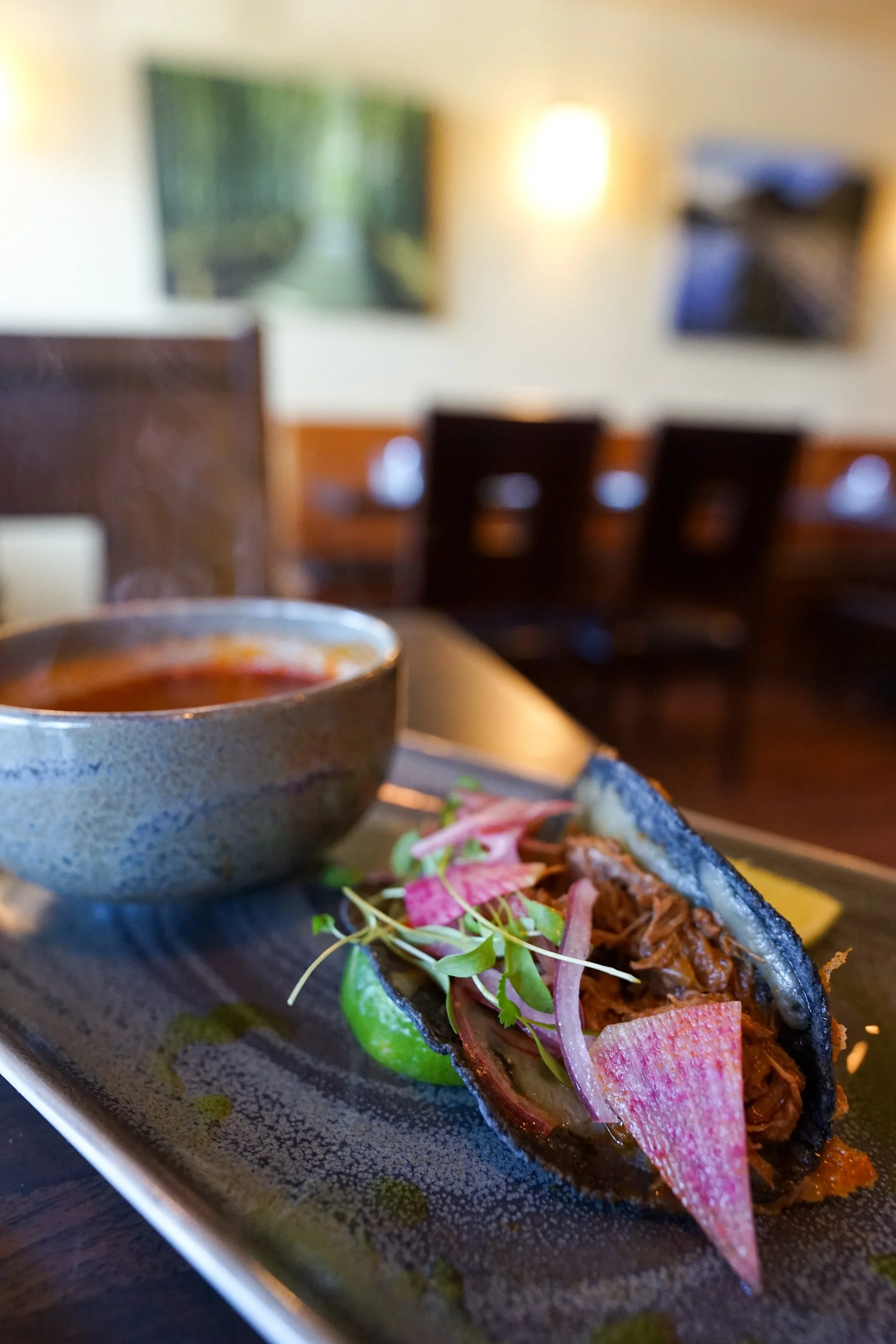 Close-up of a fish taco topped with pickled red onions, microgreens, and a wedge of lime, served on a black plate with a small cup of sauce in the background.