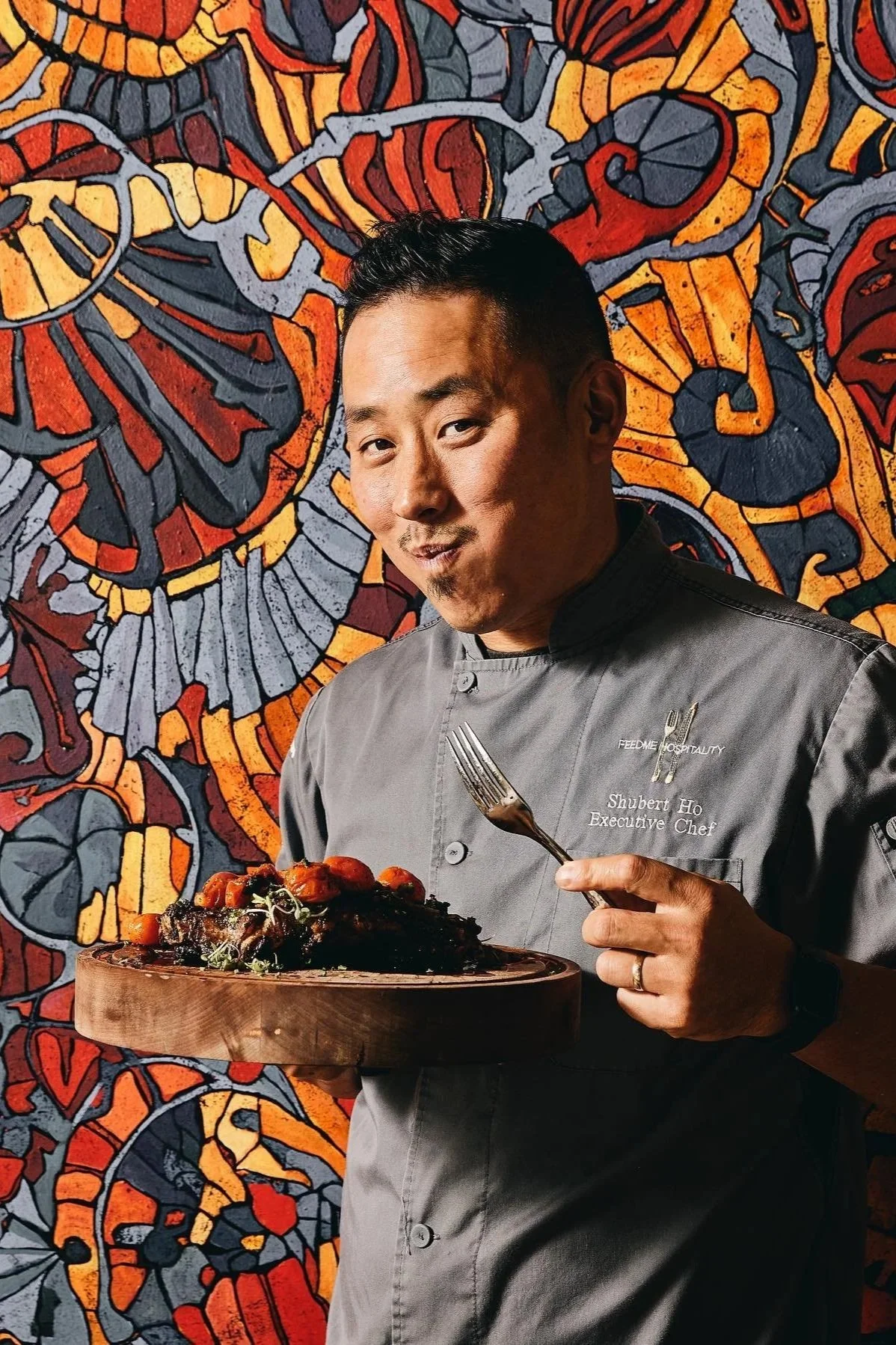 A male chef with black hair and a goatee, wearing a gray chef coat with the name 'Shubert Ho,' holding a fork and a plate of food, standing in front of a colorful, abstract mural with orange, red, gray, and black patterns.