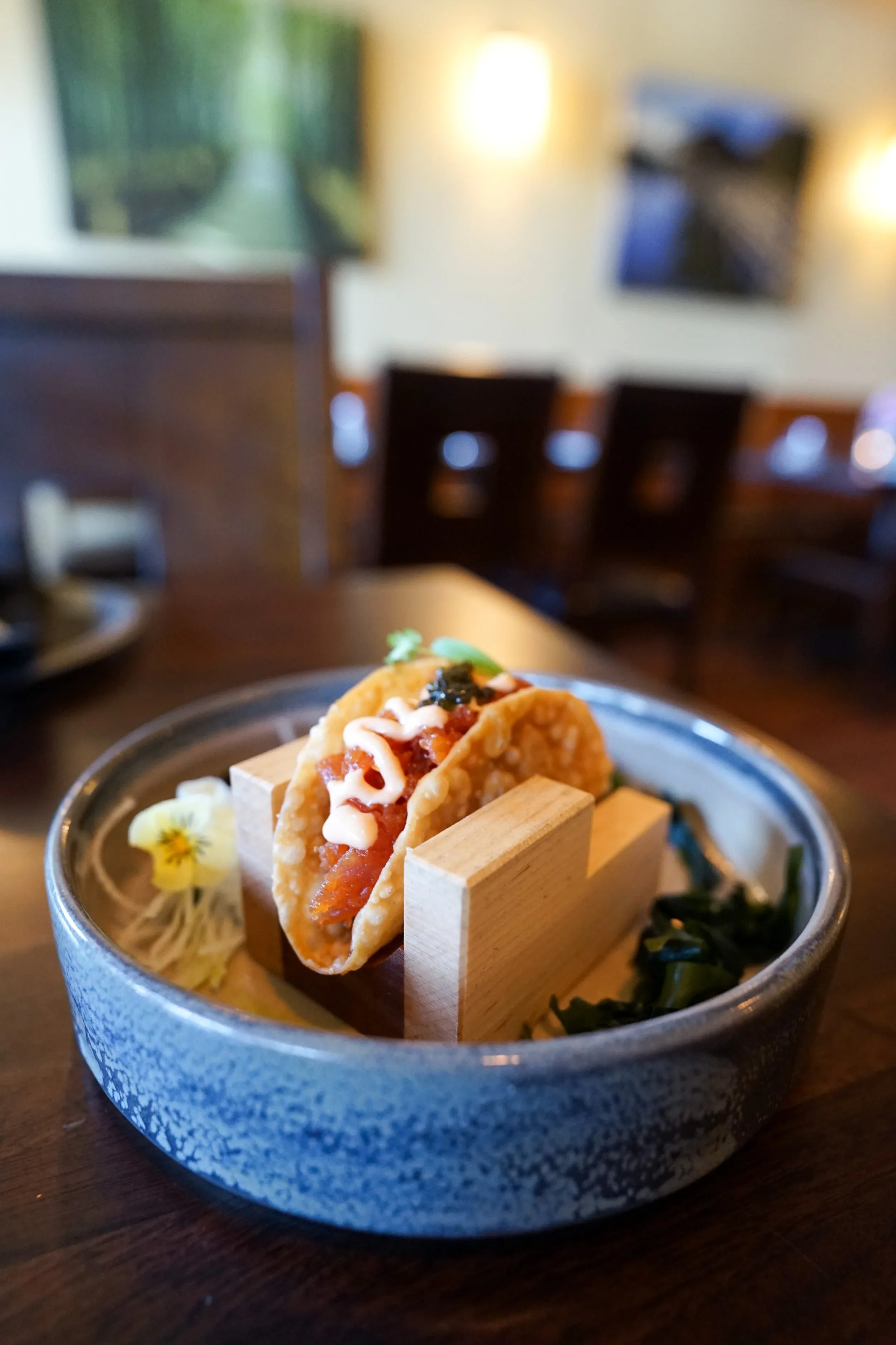Close-up of a single taco with toppings, served in a decorative bowl on a wooden table, with a blurred restaurant interior in the background.