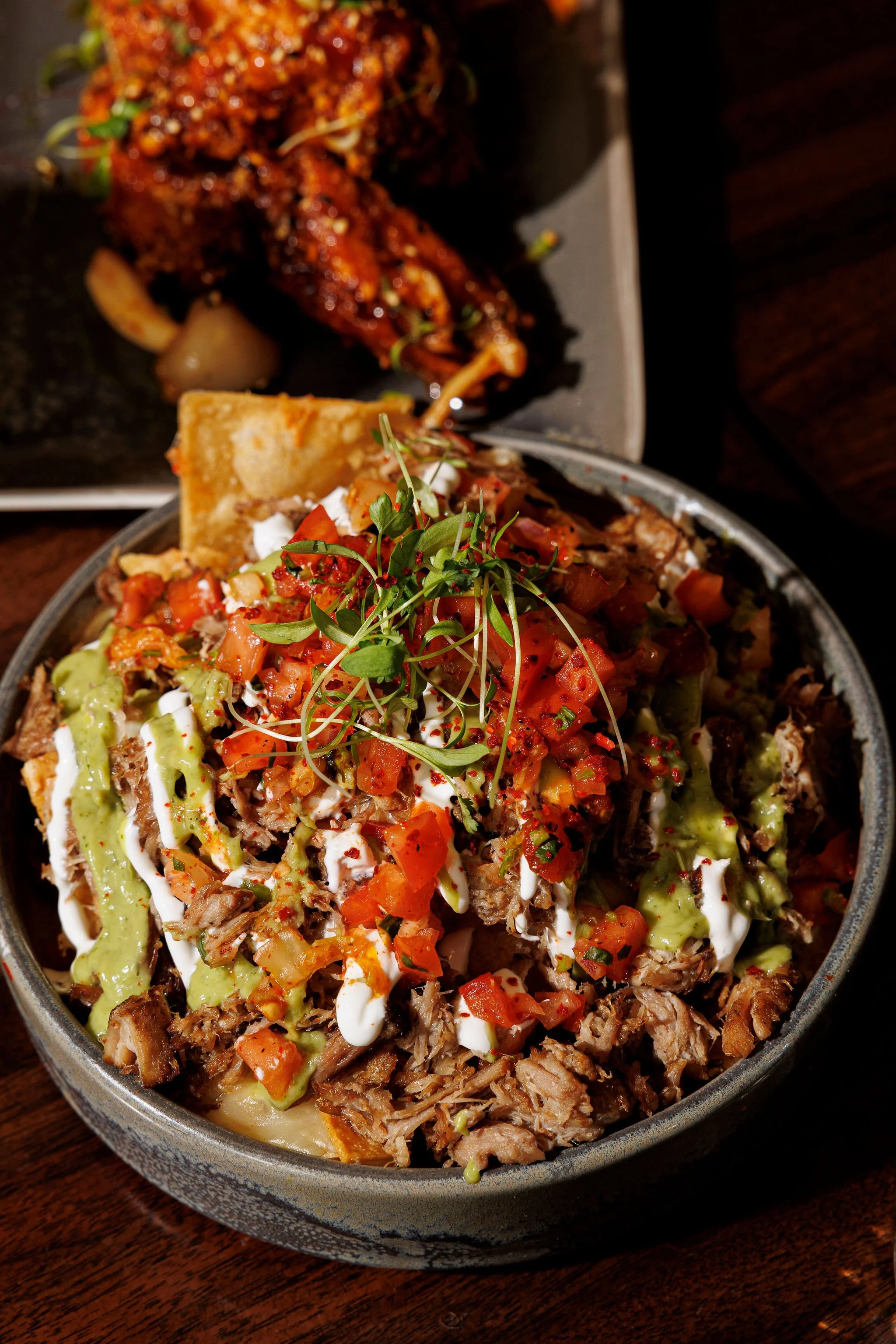 Close-up of a bowl of Mexican food topped with diced tomatoes, green sauce, white sauce, microgreens, shredded meat, and chips, with a plate of spicy chicken in the background.