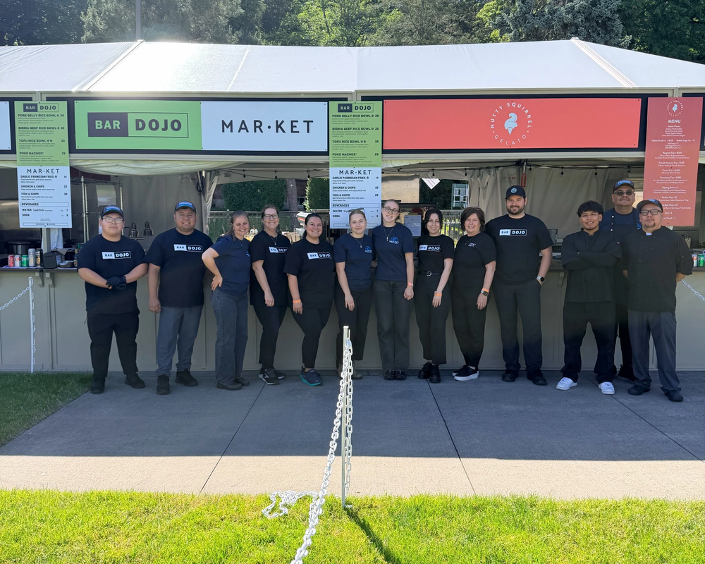 Group of 11 people standing in front of a food stall labeled 'Bar Dojo Market' at an outdoor event.