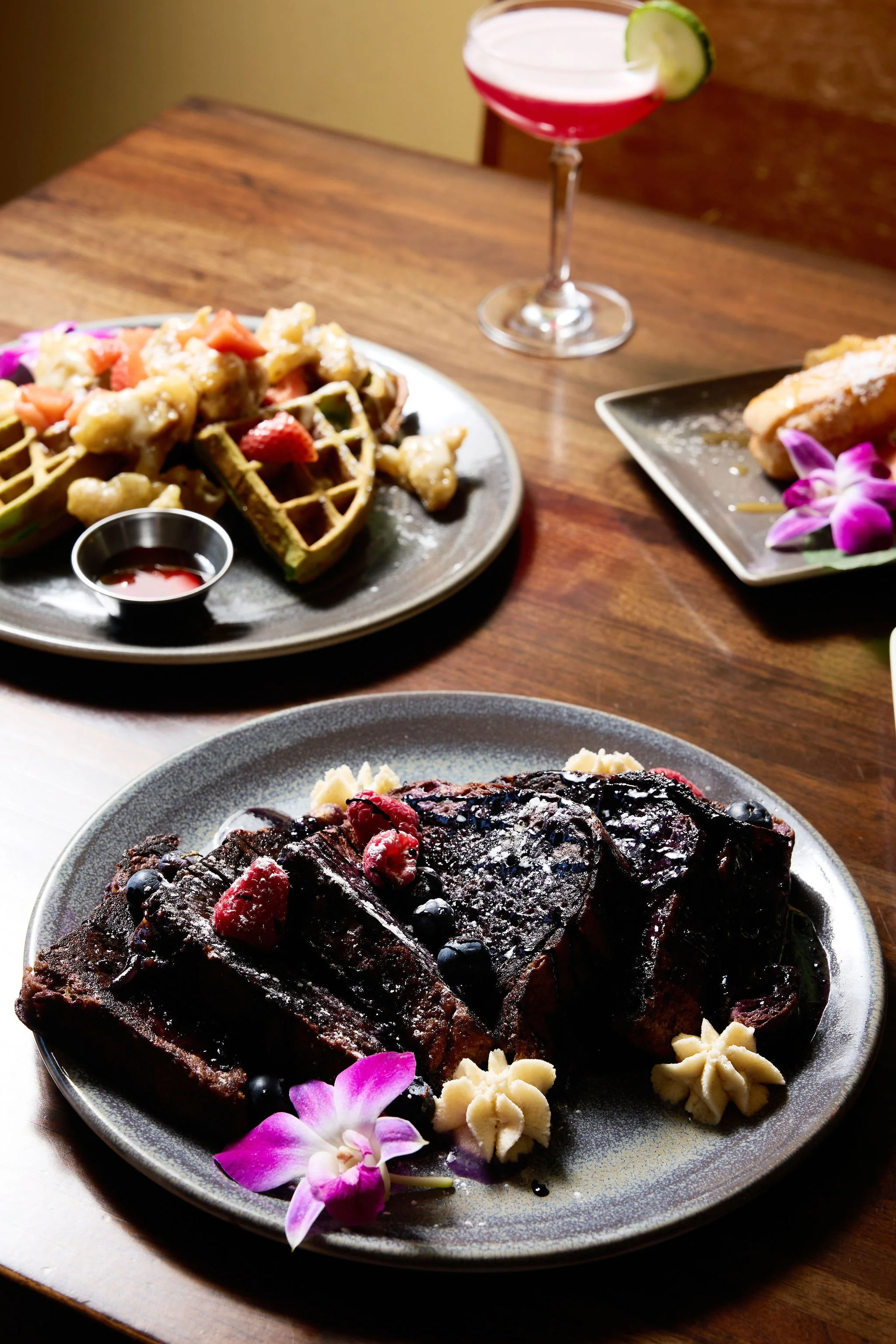 A plate of chocolate cake topped with berries and garnished with orchids and edible decorations, with a glass of red cocktail and another dessert on a table.