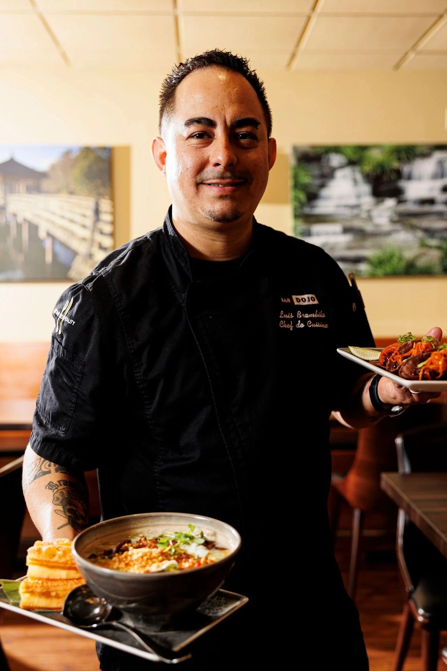 Chef in black uniform holding a plate of Asian cuisine in a restaurant. Background has framed artwork and wooden furniture.