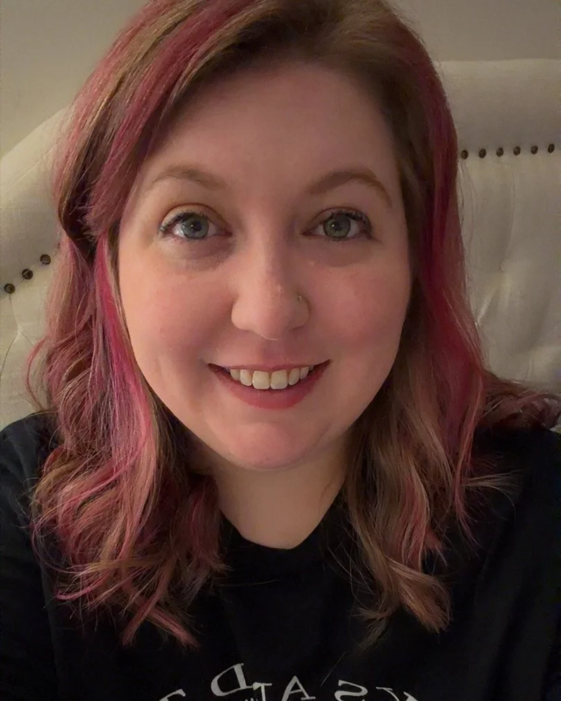 A young woman with shoulder-length wavy hair dyed pink, smiling and wearing a black shirt, sitting on a beige upholstered chair.