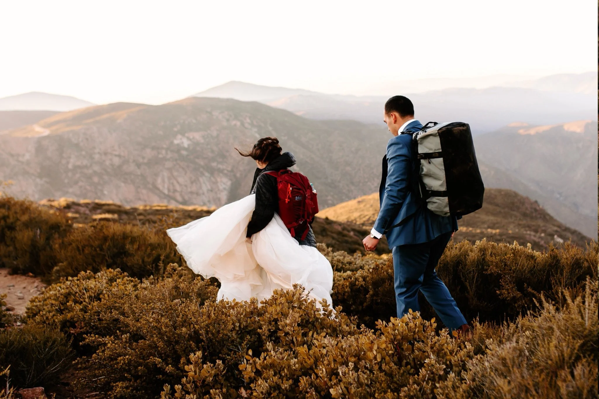 Two individuals hiking on a mountain trail with scenic mountain views in the background, one wearing a red backpack and the other wearing a large black backpack, during sunset or sunrise.