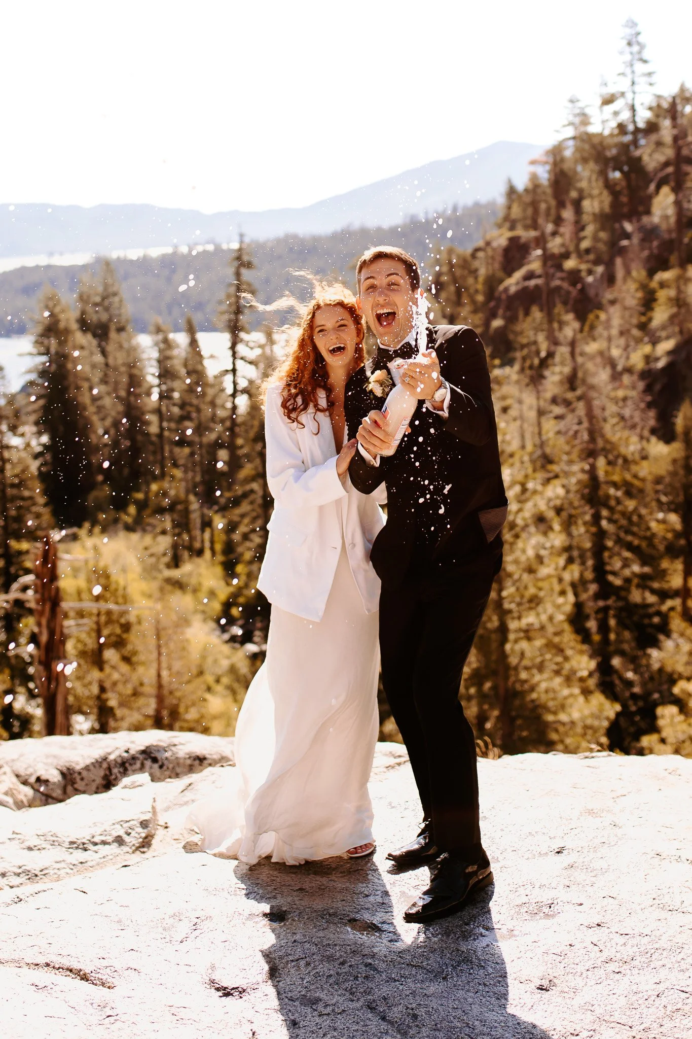 A newlywed couple celebrating outdoors in a snowy mountain landscape, with the groom spraying champagne and both smiling joyfully.