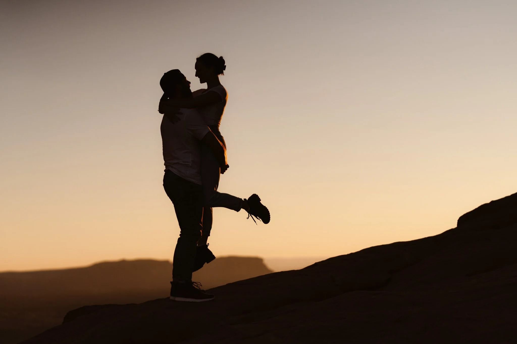 Two people, one holding the other up in a hug, silhouetted against a sunset or sunrise in a mountainous landscape.