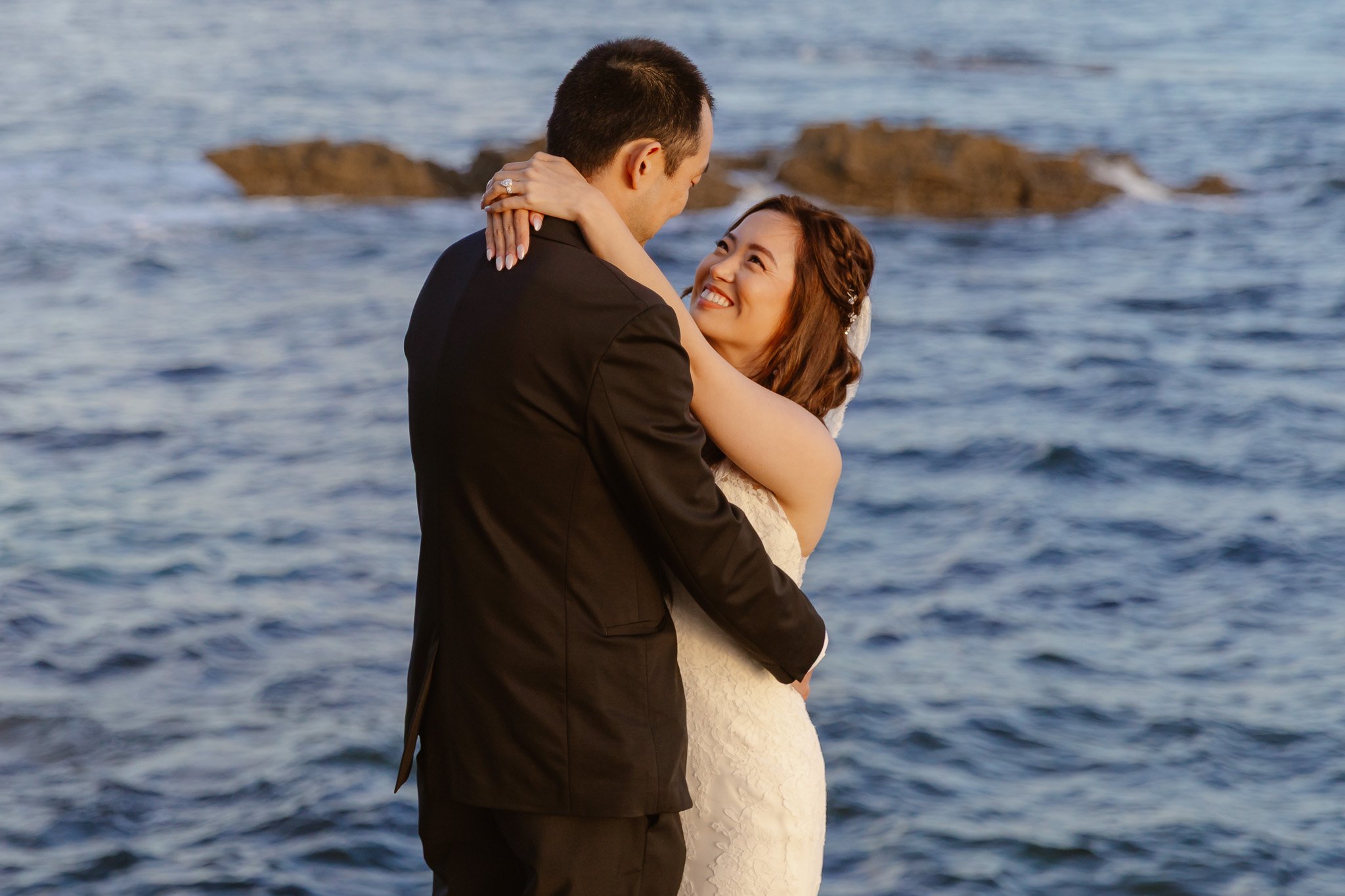 A man in a black suit holding a woman in a wedding dress near the water, with rocks in the background.