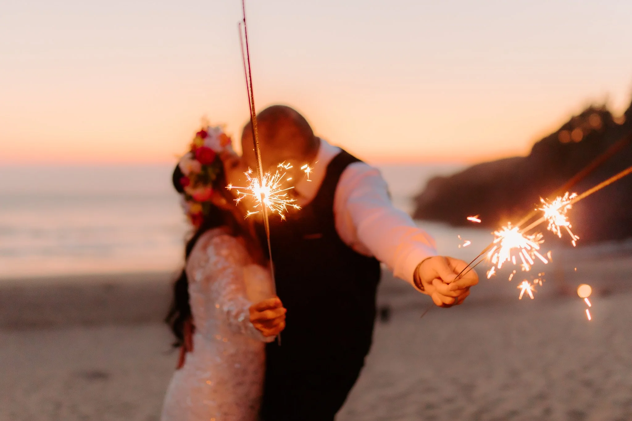 Two people holding sparklers on a beach during sunset, with the sky in pastel colors and a person wearing a floral headpiece.
