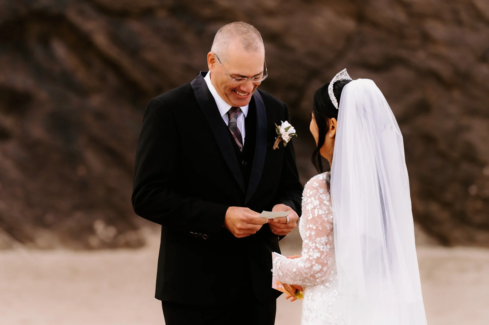A groom in a black suit with glasses and a boutonniere is smiling as he exchanges vows with a bride in a white lace dress and veil surrounded by a blurry outdoor natural background.