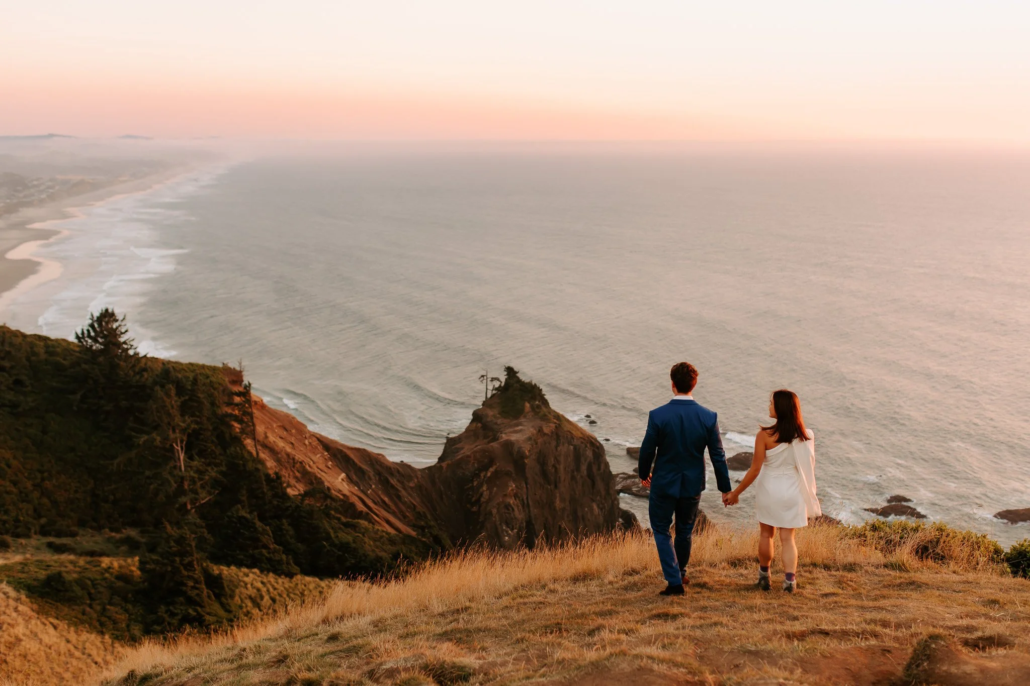 A man and woman holding hands and walking along a grassy hillside overlooking the ocean at sunset. The man is wearing a blue suit and the woman is wearing a white dress.