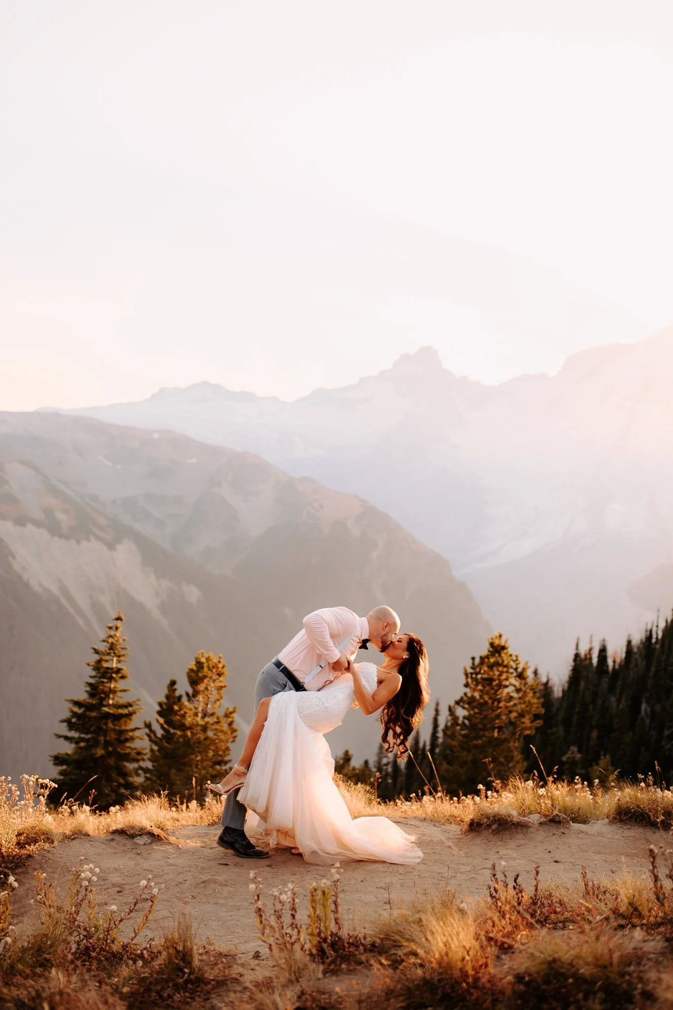 A couple dancing outdoors in a mountainous area with trees and wildflowers, with the man dipping the woman as they kiss during sunset.