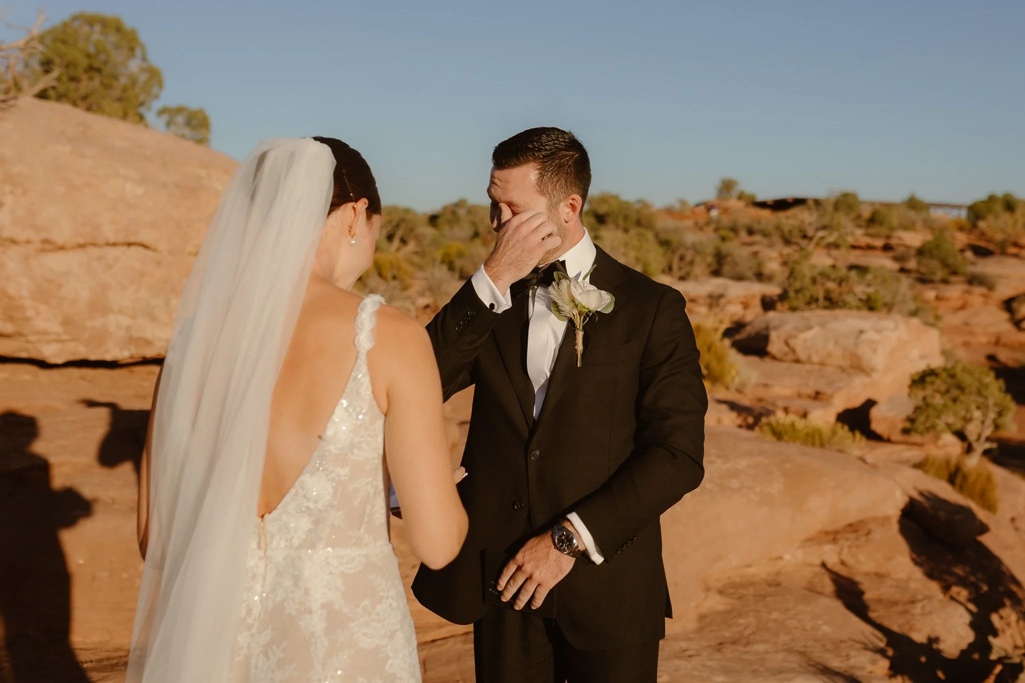 A bride and groom are shown during their wedding ceremony outdoors in a desert landscape with rocky terrain and shrubs, under clear blue skies.