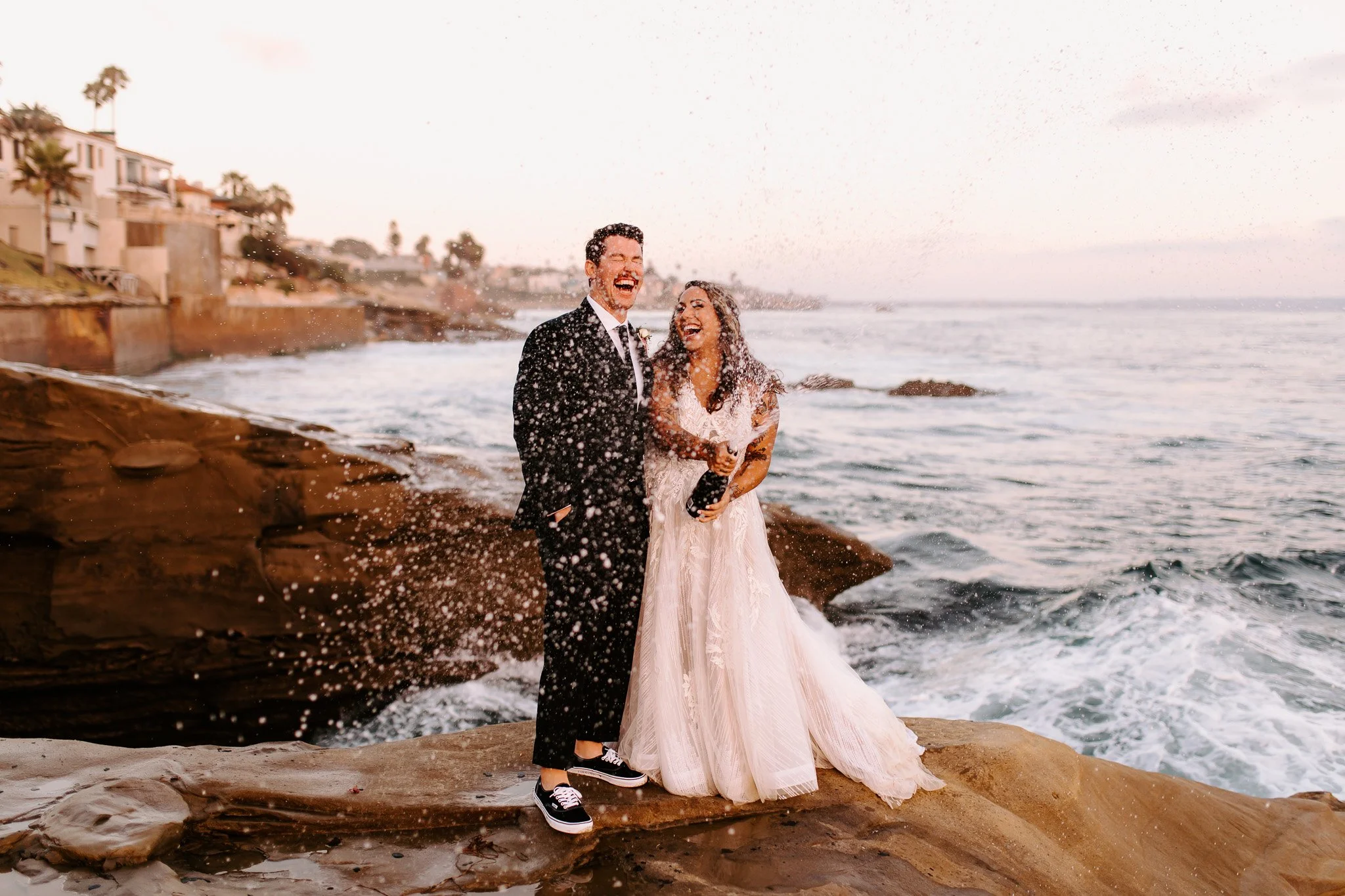 A newlywed couple dressed in wedding attire standing on rocky beach near the ocean, smiling and laughing as water splashes around them.