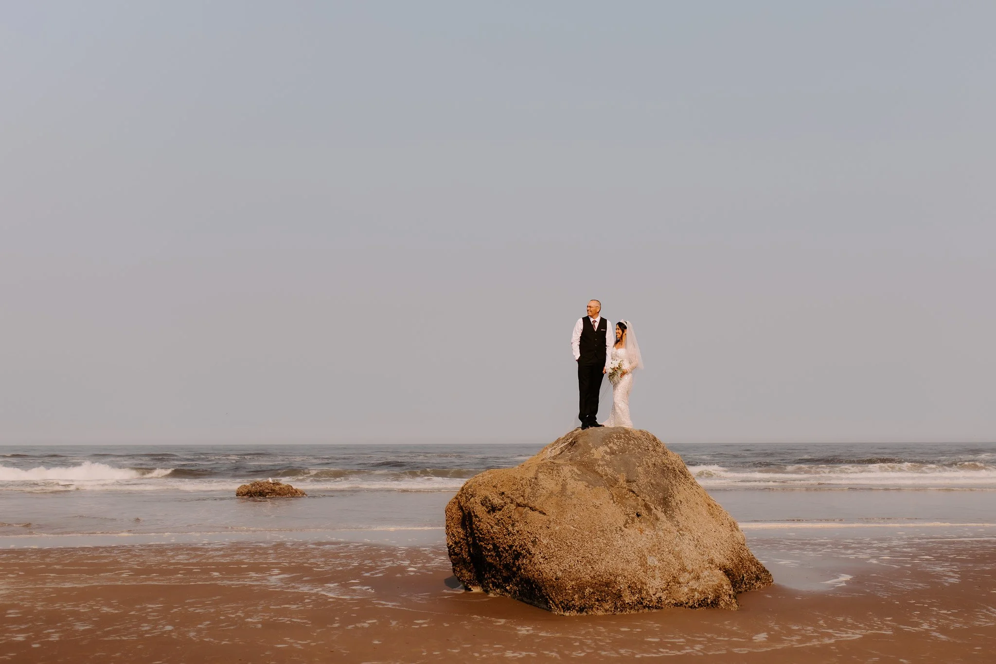 A bride and groom standing together on a large rock at the beach during sunset, with ocean waves in the background.