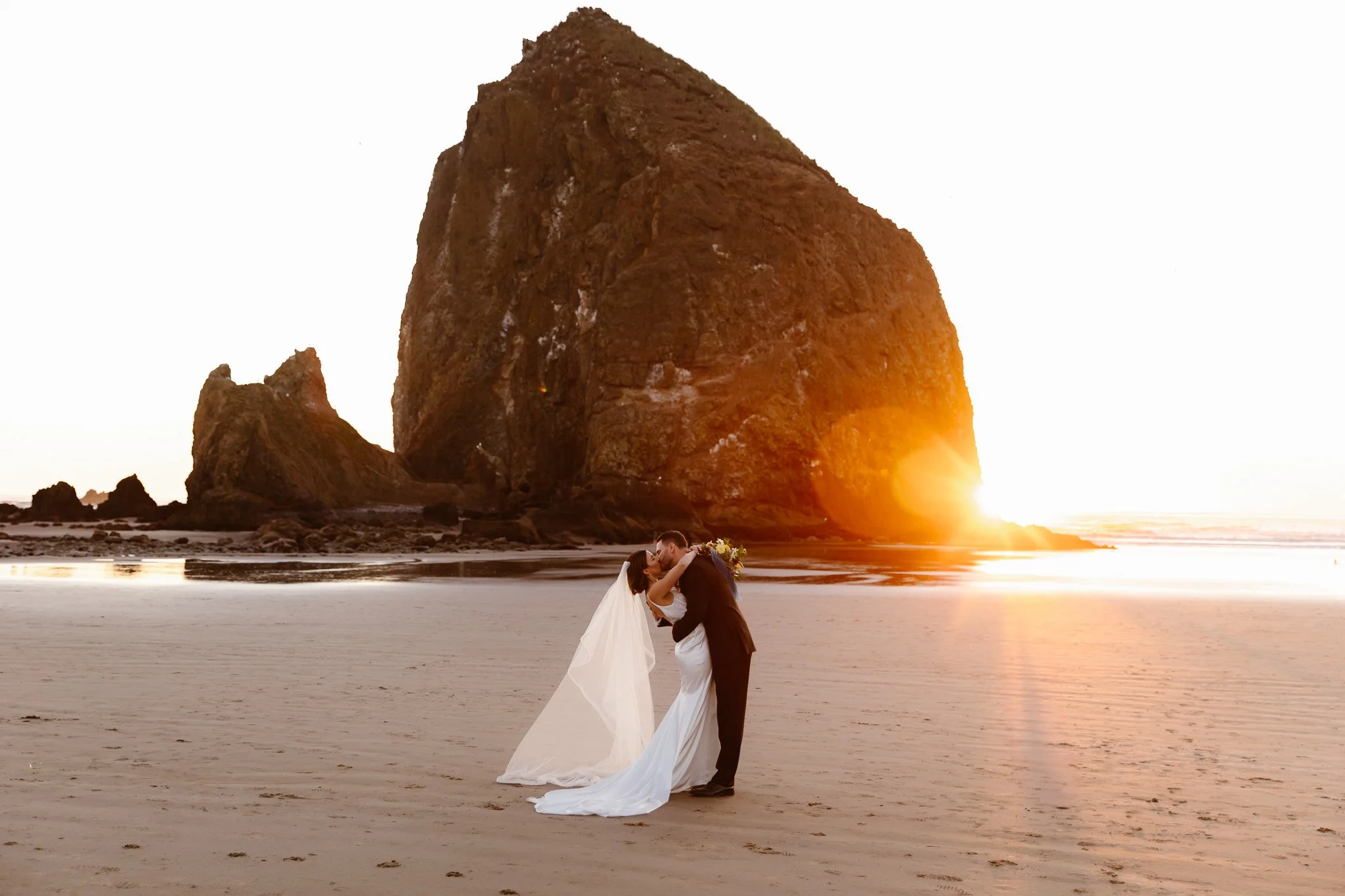 A bride and groom kiss on a beach at sunset with a large rock formation in the background.