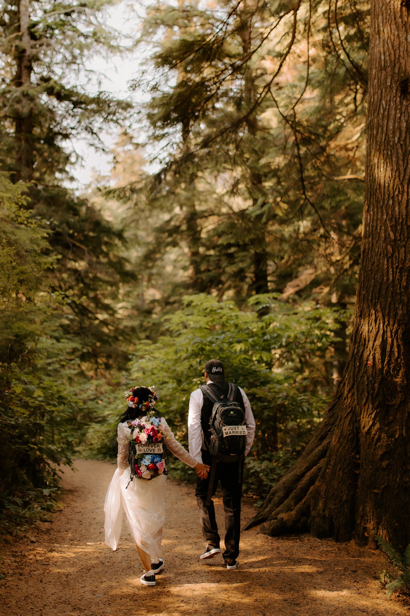 A bride and groom walking hand in hand through a forest. The bride is wearing a wedding dress covered with a floral shawl, and the groom is wearing a hat. They are holding hands and have signs on their backs that read 'Forever Love' and 'Just Married