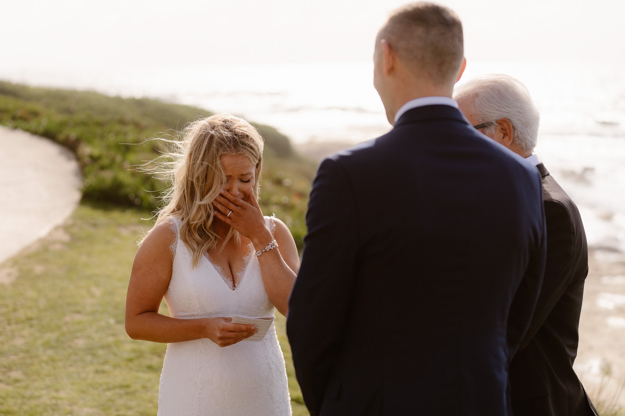 A bride in a white wedding dress crying during her wedding ceremony on the beach, with a man in a dark suit facing her and an older man with white hair behind him.