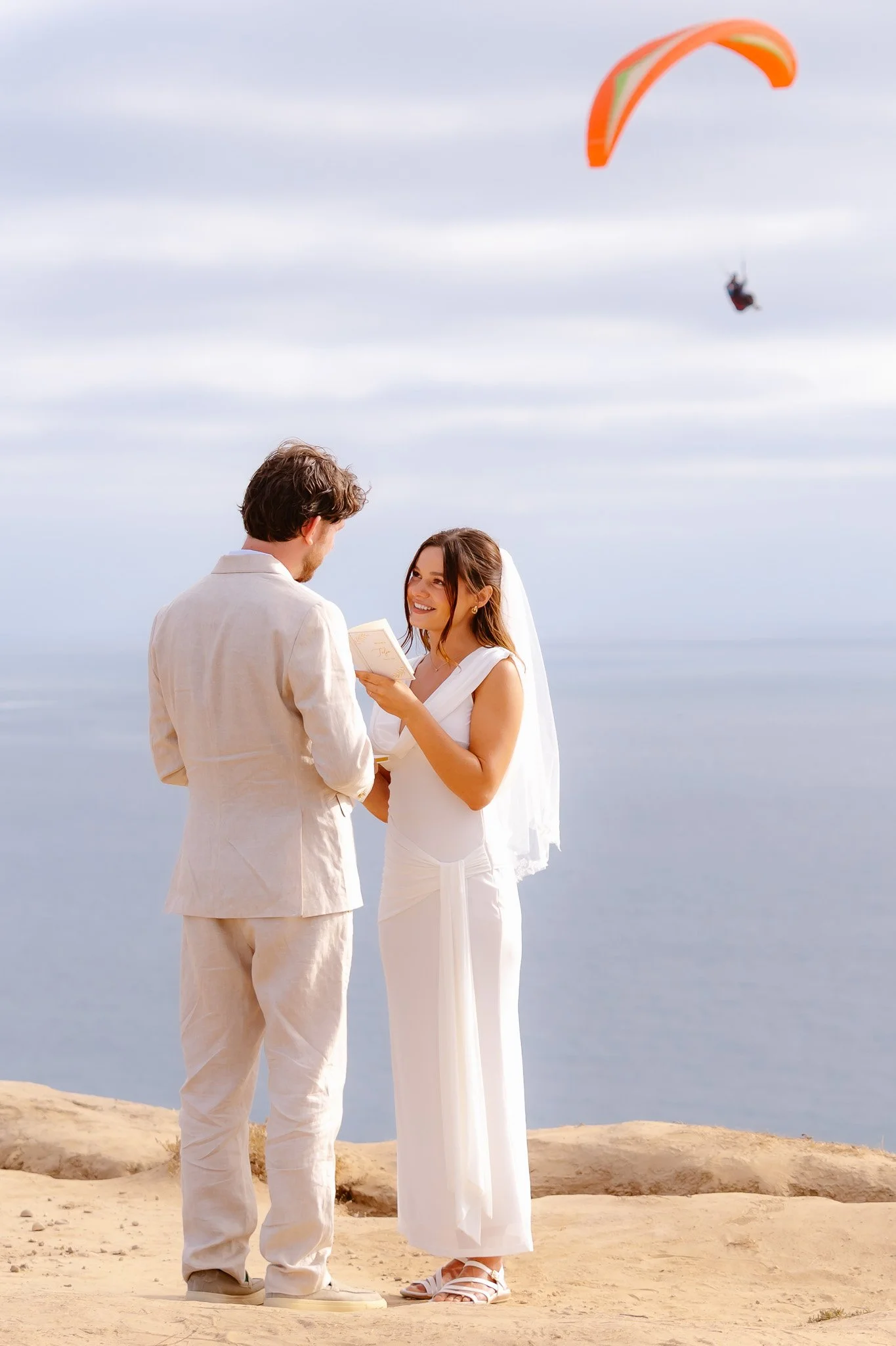 A couple dressed in white formal clothing stand on a sandy beach with a body of water in the background. They are exchanging vows, with the woman smiling while holding a small book, and the man facing her. An orange and white kite is flying in the sky above them.