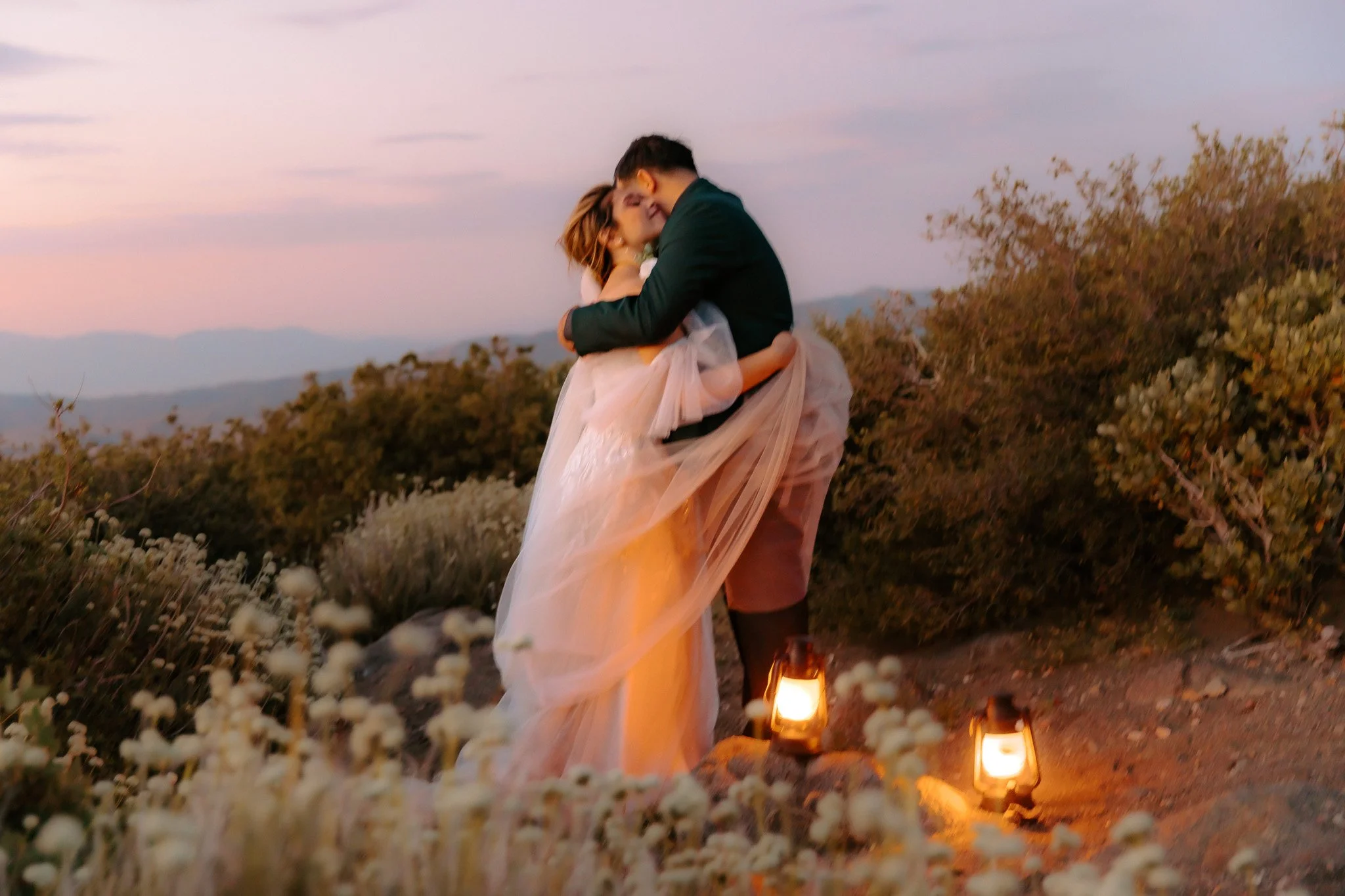 A couple, dressed in wedding attire, sharing a kiss outdoors during sunset, surrounded by flowers and lanterns.