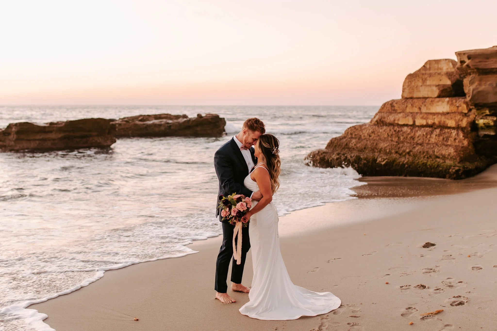 A couple dressed in wedding attire standing closely on a beach at sunset, with cliffs in the background. The bride is holding a bouquet of pink and white flowers.