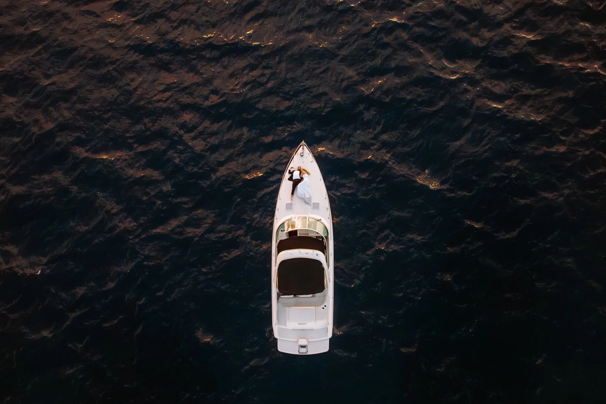 An aerial view of a yacht with two people lying on the deck, floating on dark ocean water.