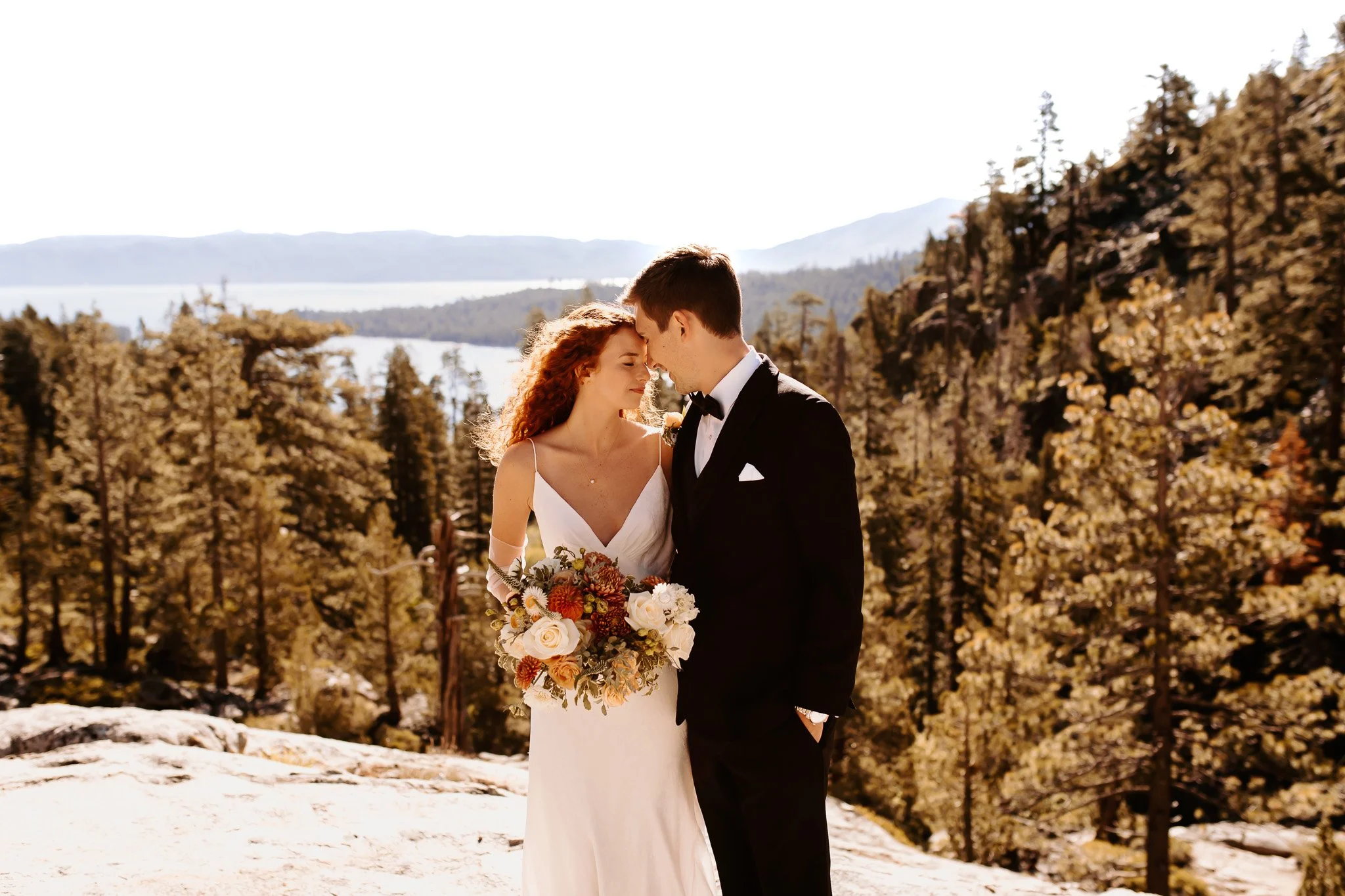A bride and groom stand close together outdoors in a forested area with mountains and a lake in the background, with their foreheads touching in an intimate moment.
