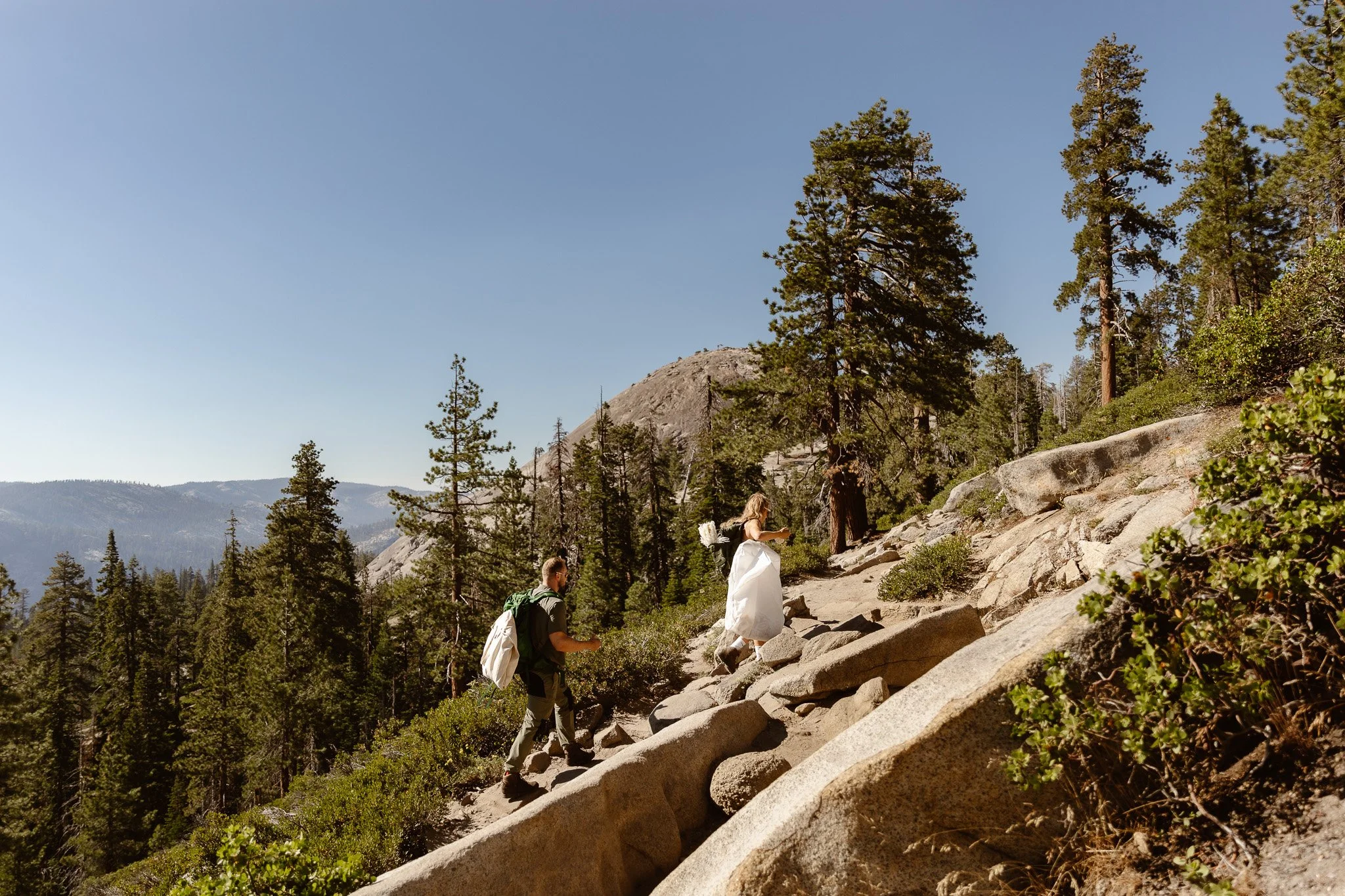 Hikers ascending a rocky trail surrounded by pine trees in a mountainous forest, with clear blue sky overhead.