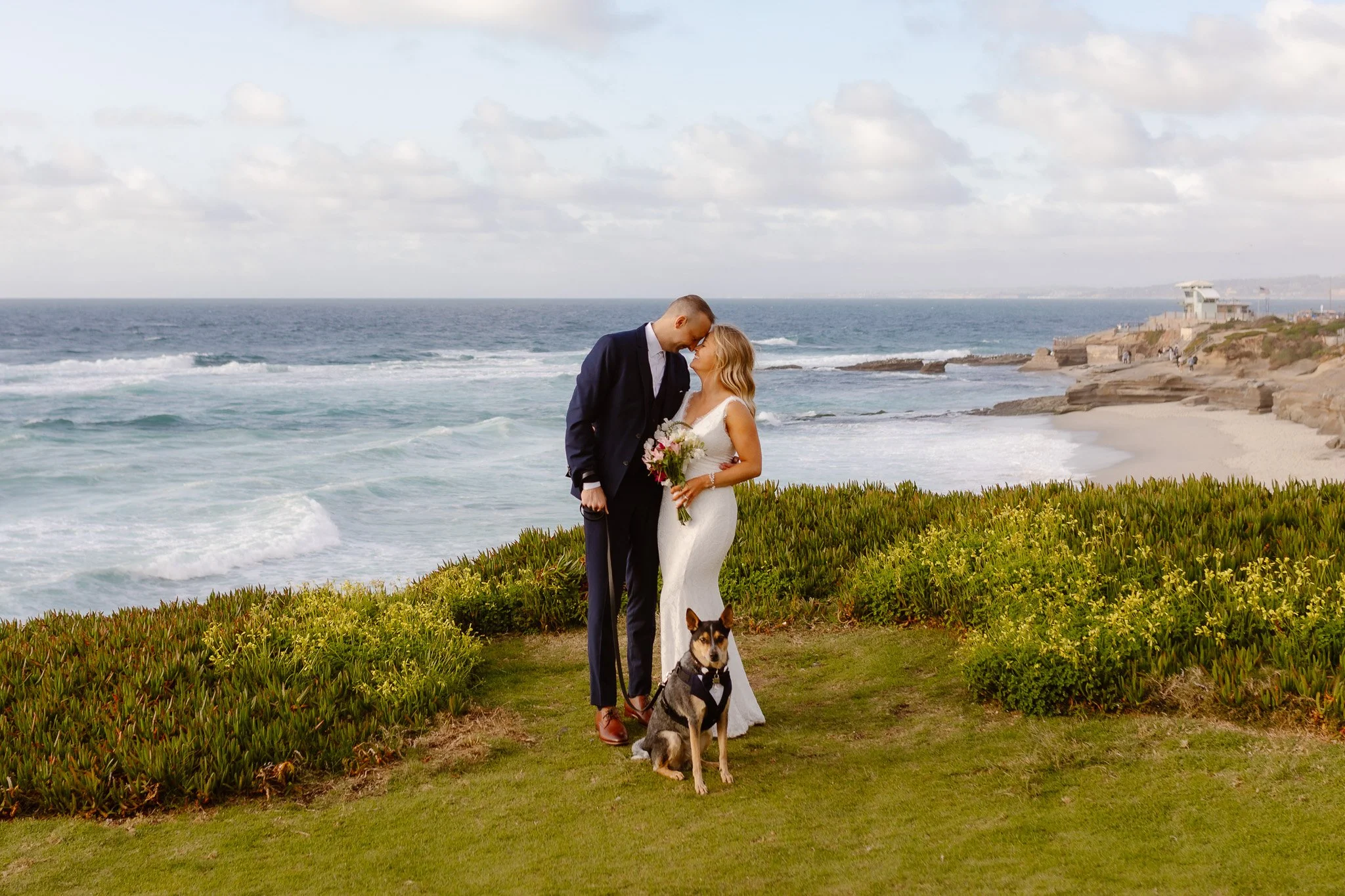 A couple dressed in wedding attire standing close by the ocean with a dog on a leash, overlooking the beach and cliffs.