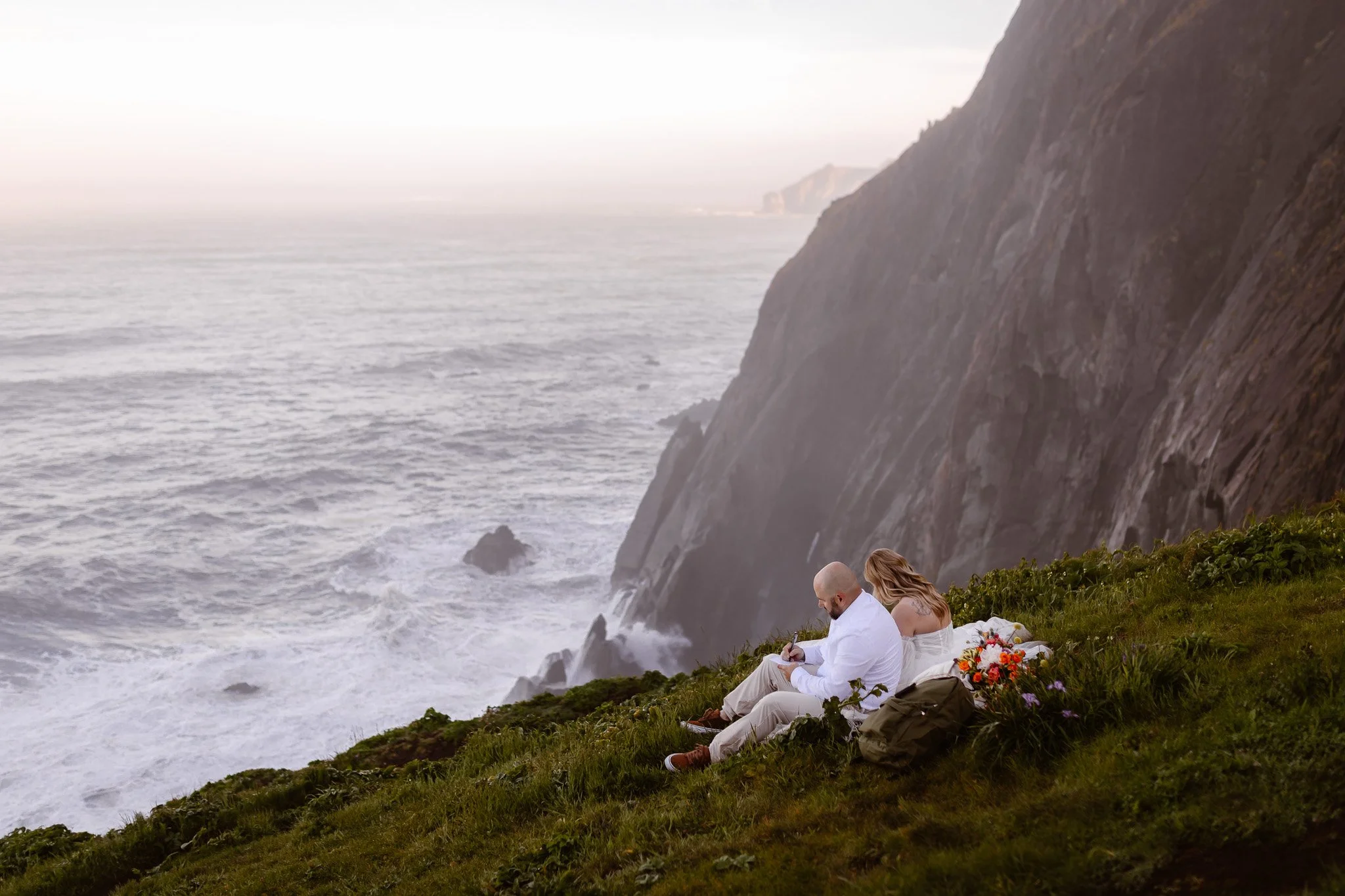 A couple sits on the grassy edge of a cliff near the ocean, with a large wave crashing below and cliffs in the distance.