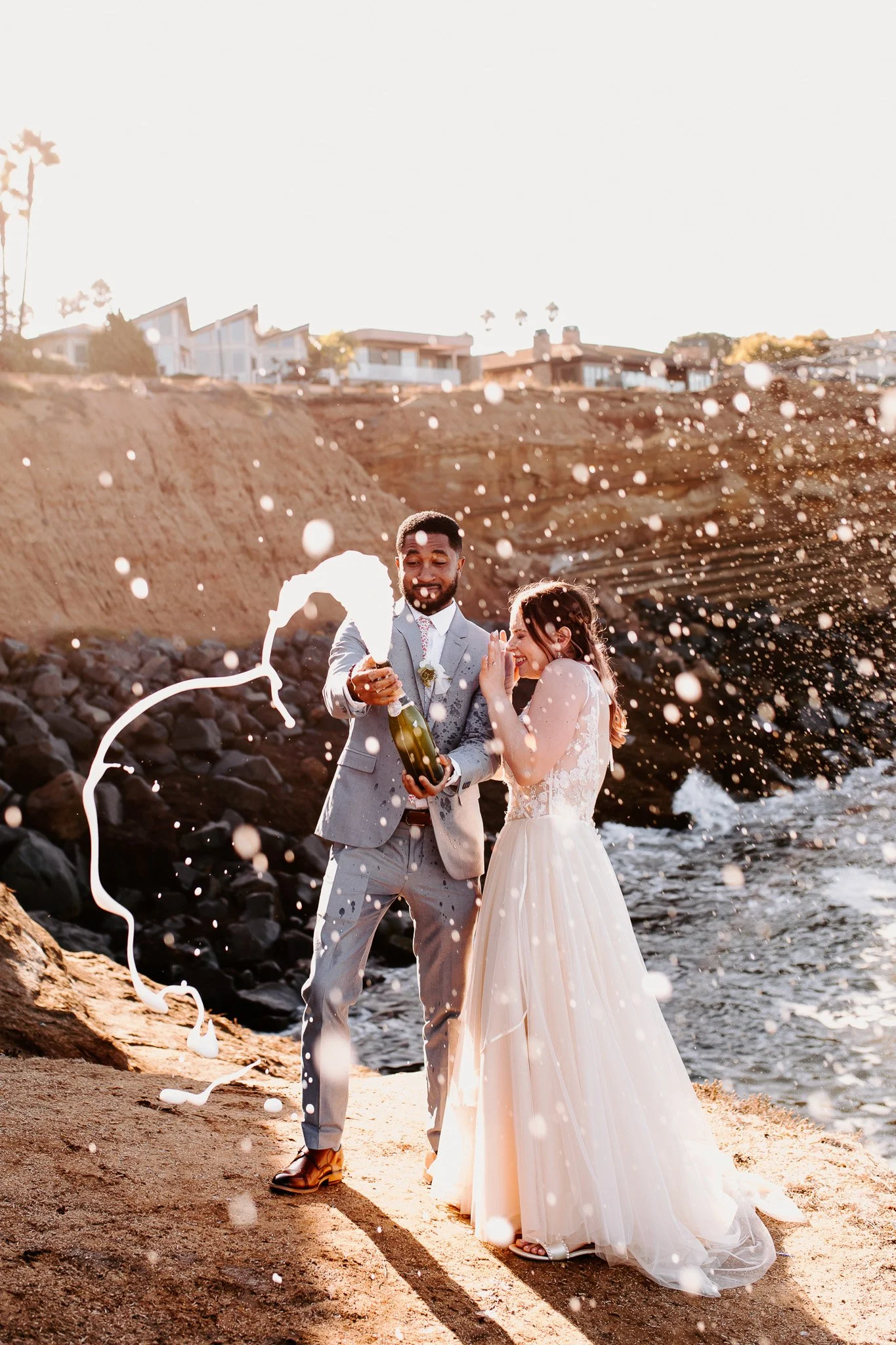 A bride and groom celebrating their wedding on a rocky beach, with water splashing around them as they open a champagne bottle and smile.